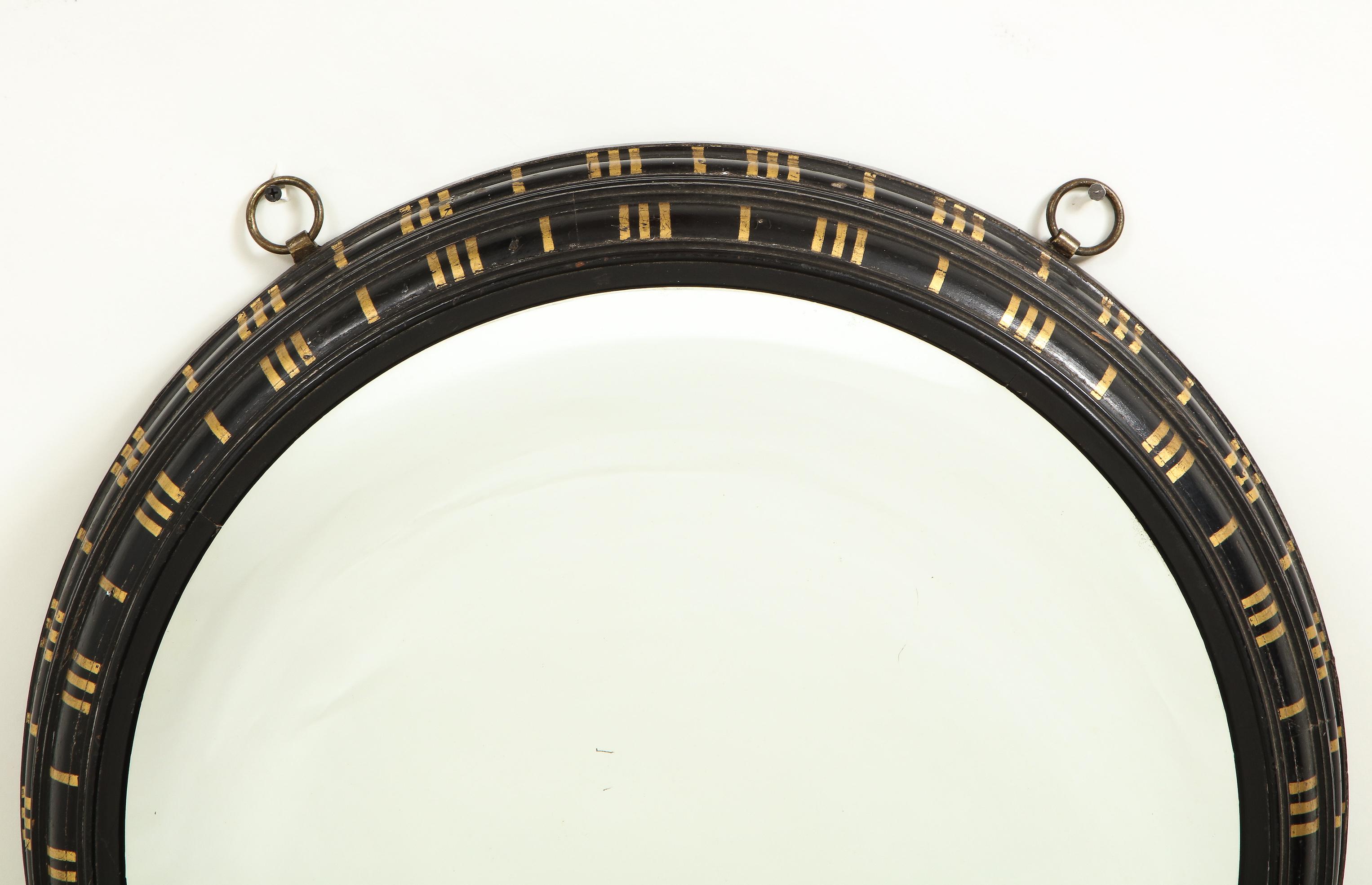 The round concave mirrored glass plate within a black frame with gilt decoration perhaps inspired by classical triglyphs. Mounted with two metal hooks for hanging.