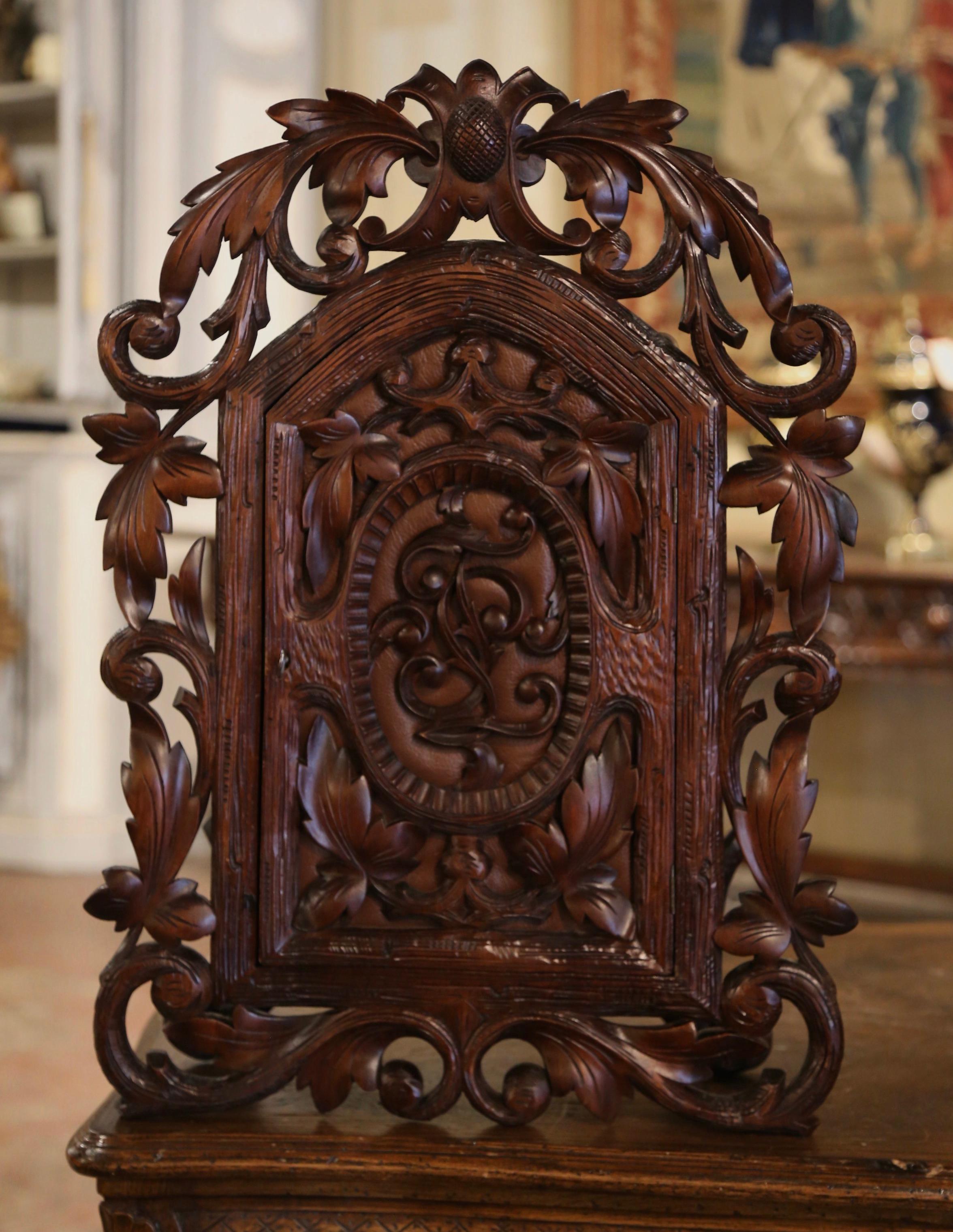 This elegant and heavily carved antique vitrine was crafted in France, circa 1880. The hanging cabinet features hand carved leaf and branch motifs throughout the front. The cabinet door with arched decor is dressed with an inset leather upholstery