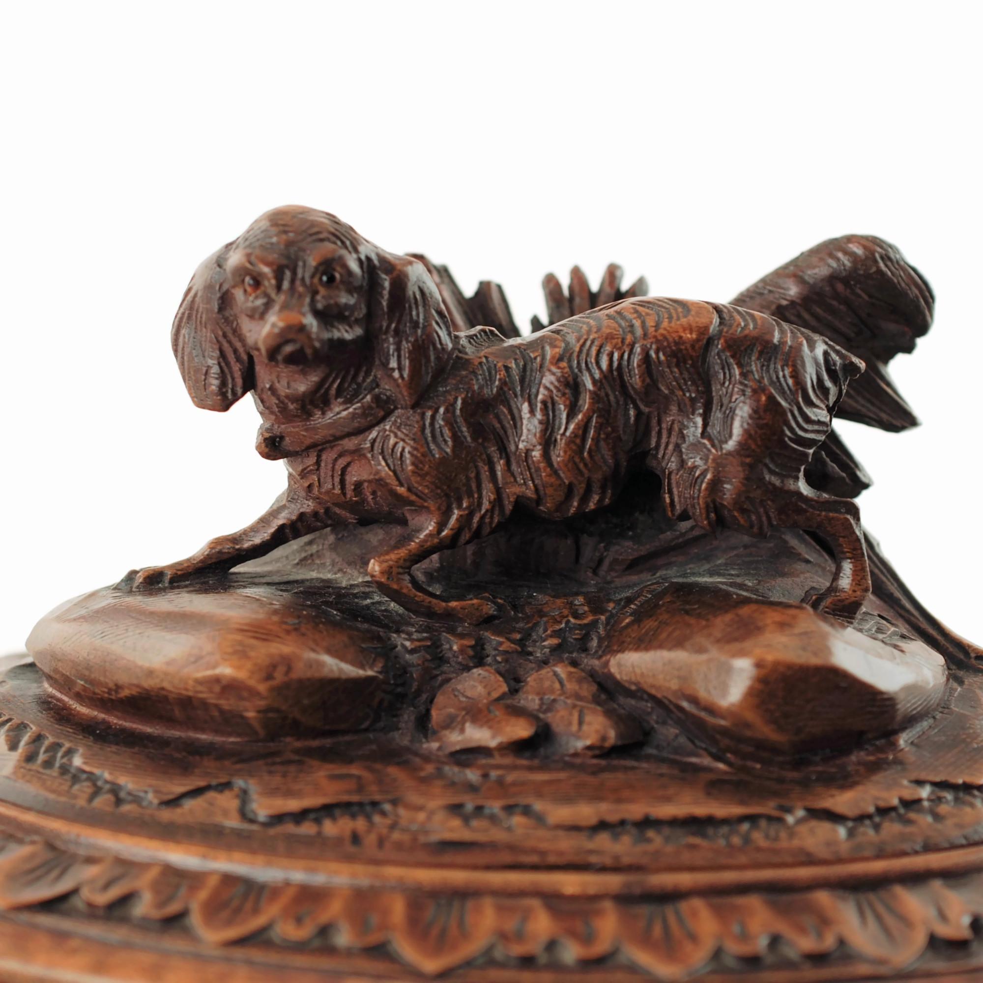 This 19th century Black Forest carved wood lockable casket has an oval shape and is topped with hinged lid featuring a fully dimensional carved spaniel with glass eyes. The dog has been depicted posed atop two rocks amongst vegetation with its mouth