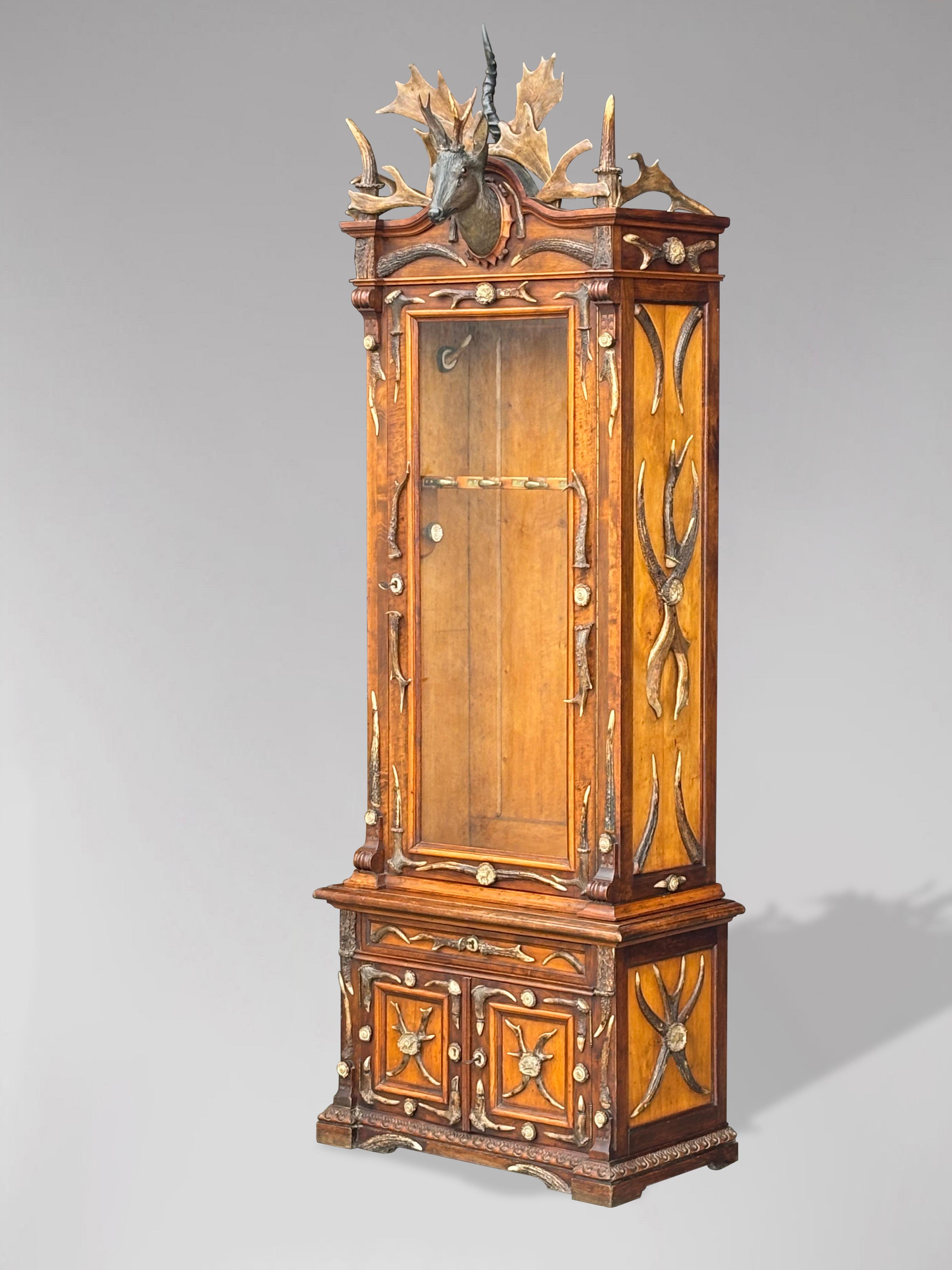 A late 19th century Black Forest, German Austrian, two parts mahogany antler gun cabinet. The top section has a single large glazed door, followed by a base with one drawer over two doors. The top is crowned with one of the most startlingly
