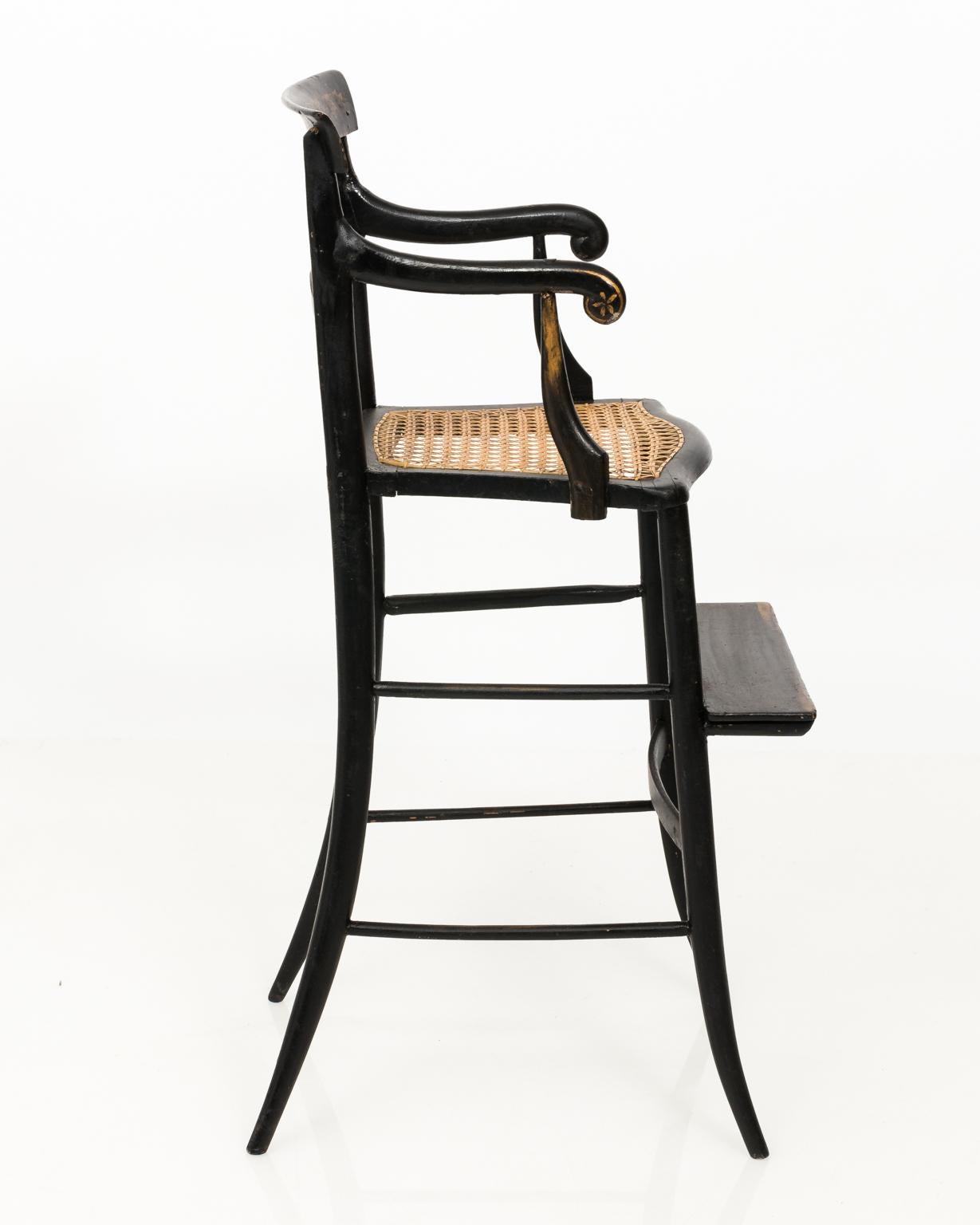 Black painted child's high chair with gold painted foliage on the seat back, scrolled arms, and a woven wicker seat, circa 19th century.