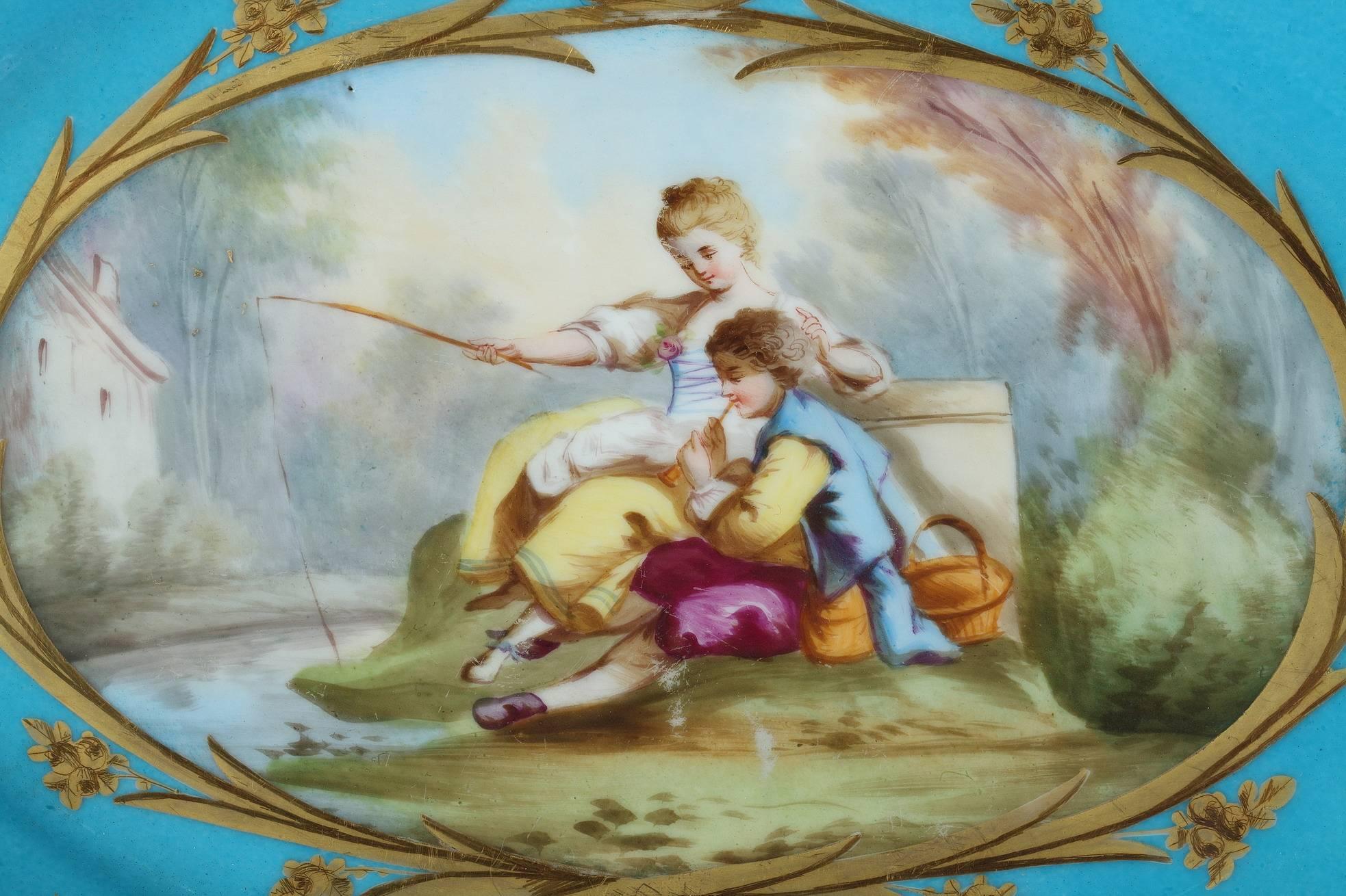 Tasse ovale en porcelaine bleue à décor polychrome composé d'un grand cartouche à bordures dorées représentant un homme et une jeune femme dans un paysage, assis sur l'herbe près d'un lac. La femme est en train de pêcher pendant que son amant joue