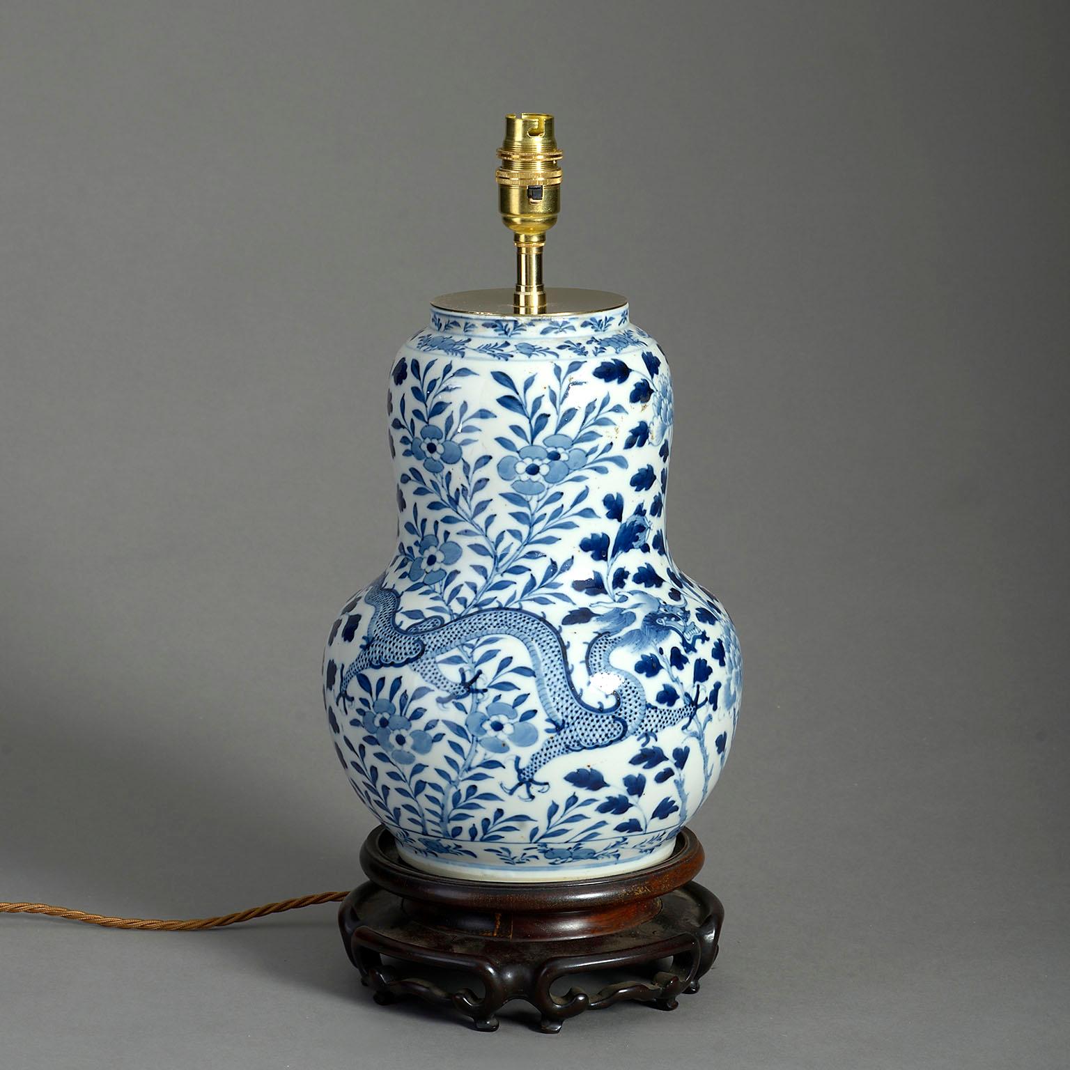 A mid-nineteenth century blue and white glazed vase of generous gourd form, decorated with dragons, flowers and foliage, set upon a carved hard wood stand and now wired as a lamp.

Dimensions refer to vase and wooden base only.

Display shade