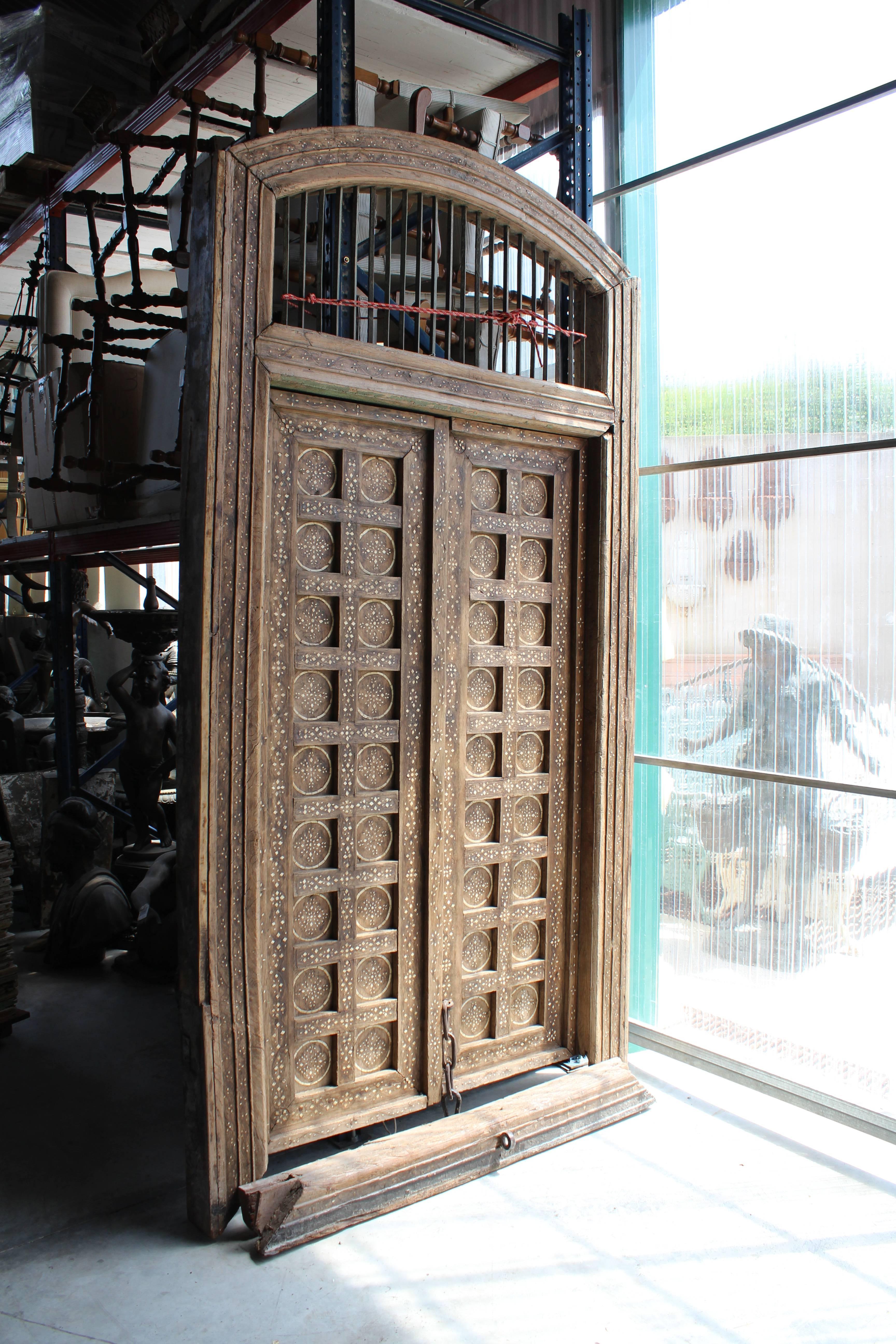 Grandiose wooden double front door with bone inlay geometric patterns decorating both panels and the frame, topped with an iron bar window.
