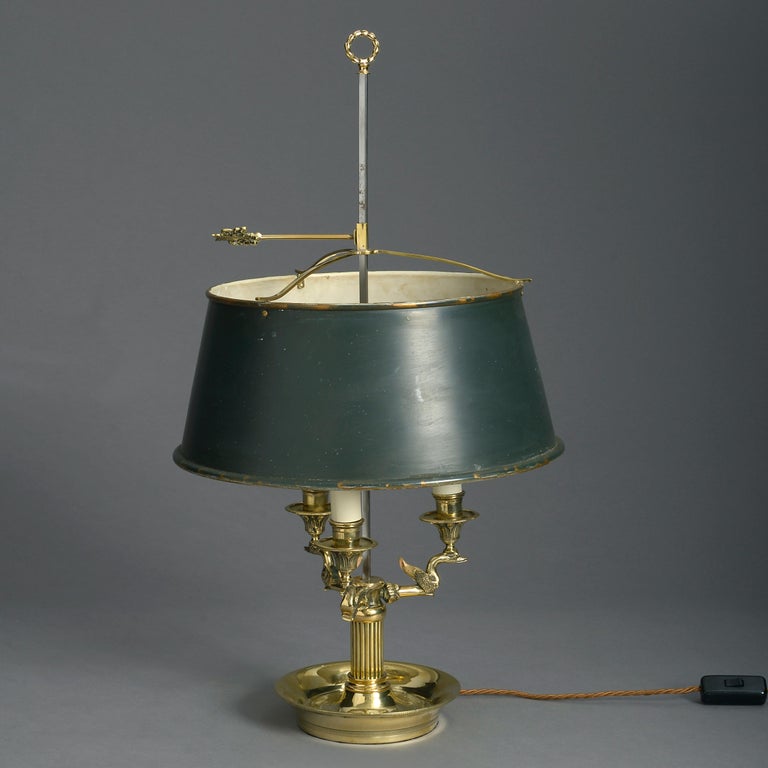 A late 19th century bouillotte lamp in the Louis XVI taste, the steel shaft with brass laurel ring and arrowhead screw, supporting a green tole shade, all above a base of three brass candle arms with stylised swan supports, set upon a fluted column