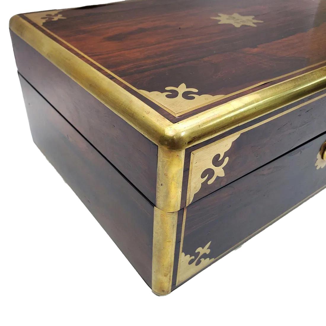 Caja inglesa de palisandro de mediados del siglo XIX con bordes redondeados de latón, la parte superior y frontal con incrustaciones de líneas de latón y dispositivos decorativos en las esquinas. El interior con una bandeja extraíble empotrada sobre