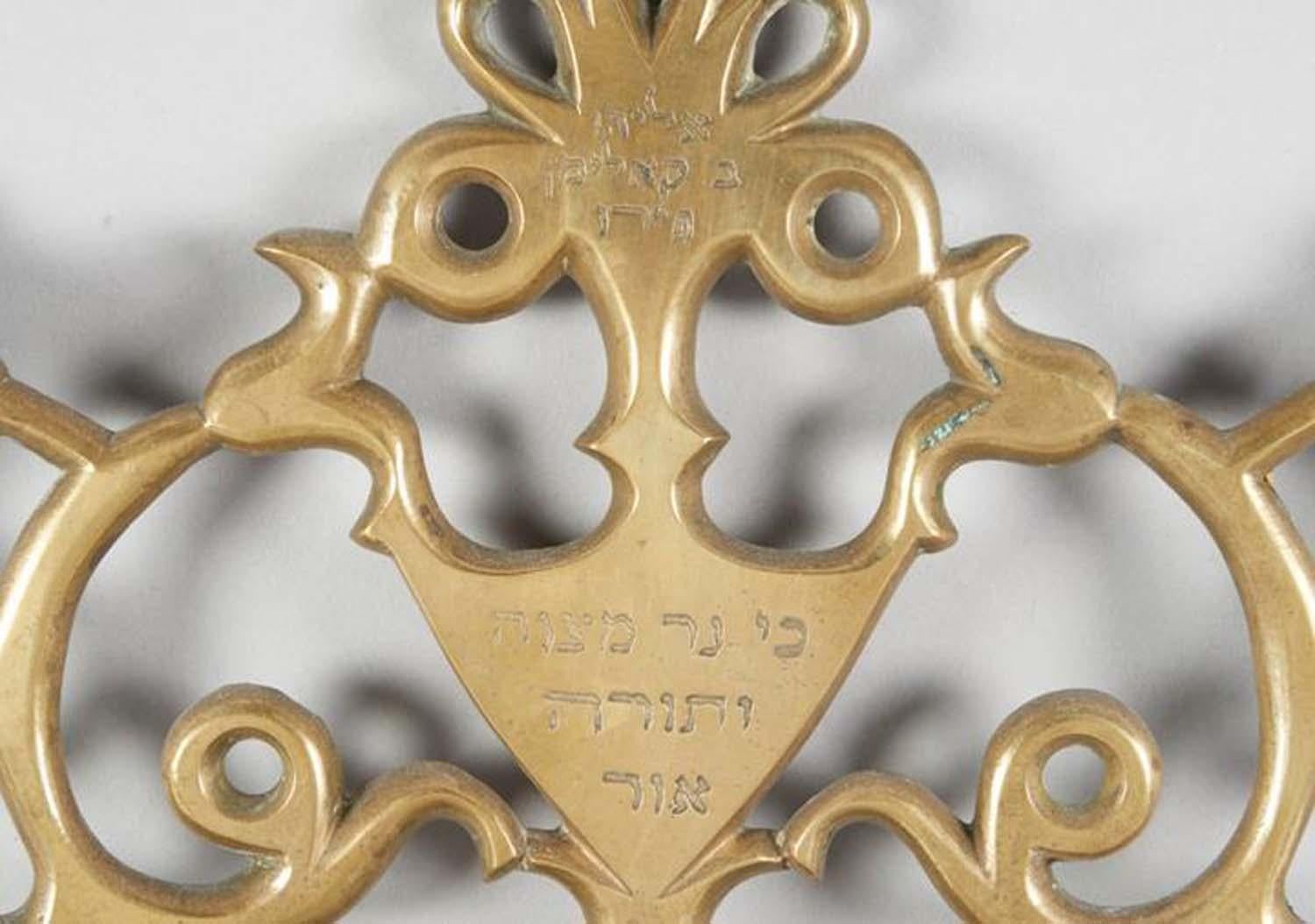 Brass Hanukkah Lamp, Salonika, Greece, 19th century.
Openwork backplate with scrolled branches; three stylized flowers support central triangular cartouche bearing Hebrew inscription “For the commandment is a lamp; the teaching is a light” (Proverbs