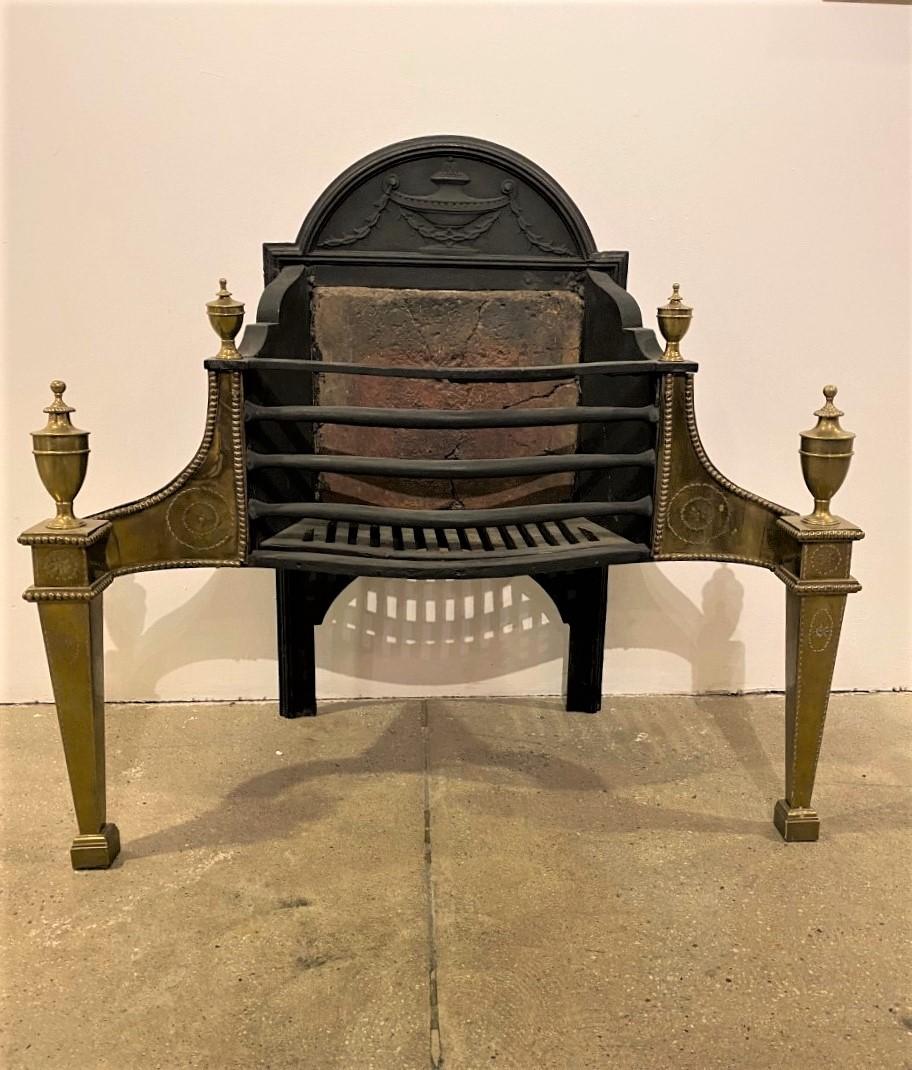 A large 19th century English Adam polished brass fire grate basket. The railed serpentine basket is set between square tapered legs with decorative embossing and beading and topped with large brass urns. wrought iron fire bars and a decorative