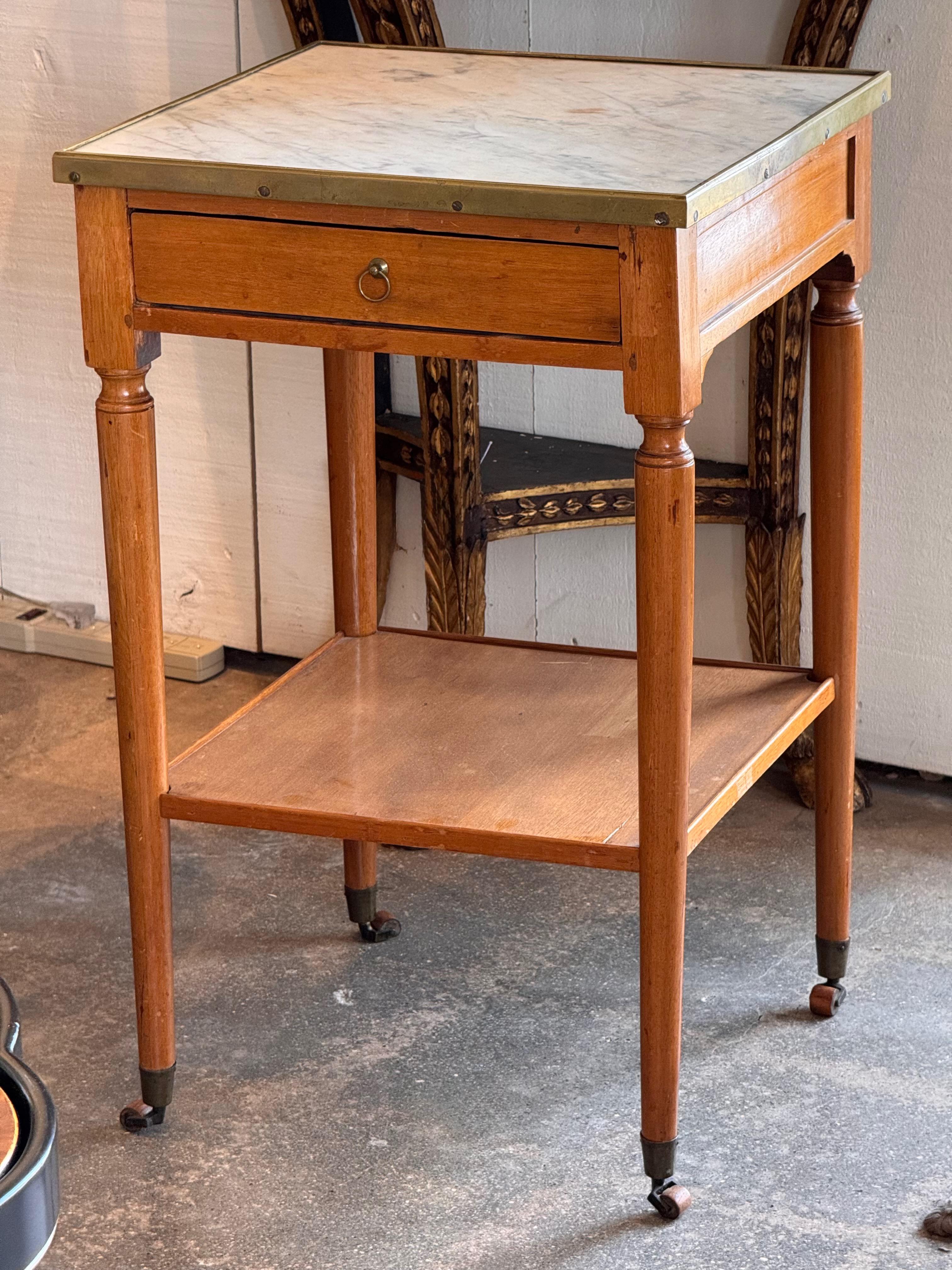 19th-century brass-mounted table with a marble top, brass trim, and a single drawer with a brass pull. The turned legs support a lower shelf for additional storage, and the piece rests on casters for easy mobility. A well-crafted, functional accent