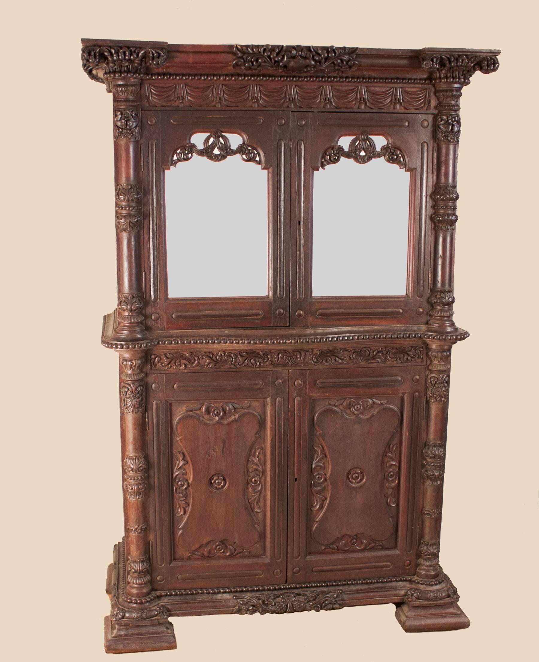 An ornately hand-carved teak wood cabinet from British India, circa 1875. In original condition, this richly colored hutch features four doors (two mirrored) and four spacious shelves inside. The piece is flanked by columns with carved capitals and