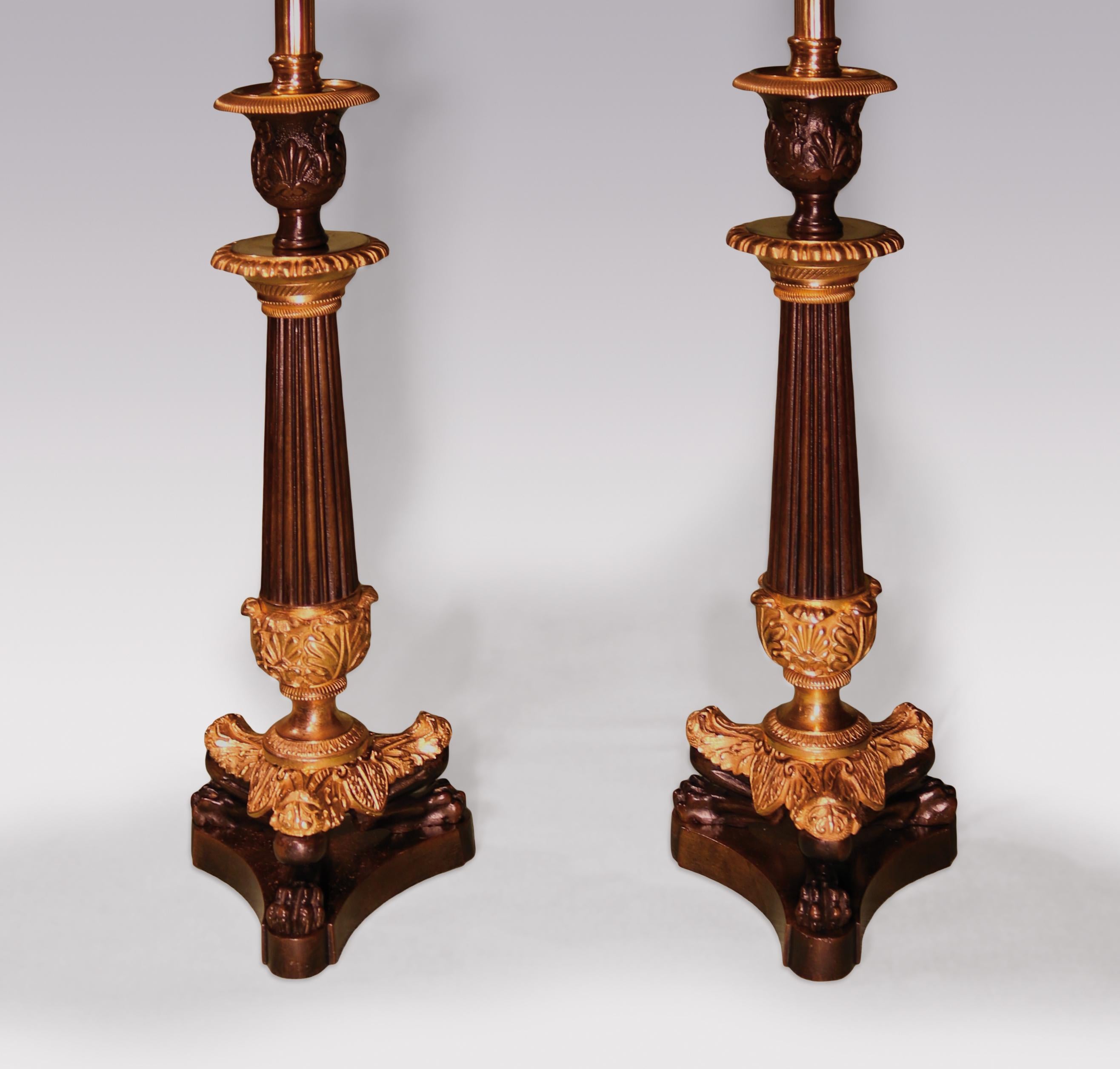 A pair of mid-19th century bronze and ormolu candlesticks, having urn-shaped sconces above floral decorated collars on reeded tapering stems, supported on acanthus decorated lion's paw triangular plinths ending on concave triform bases. (Now