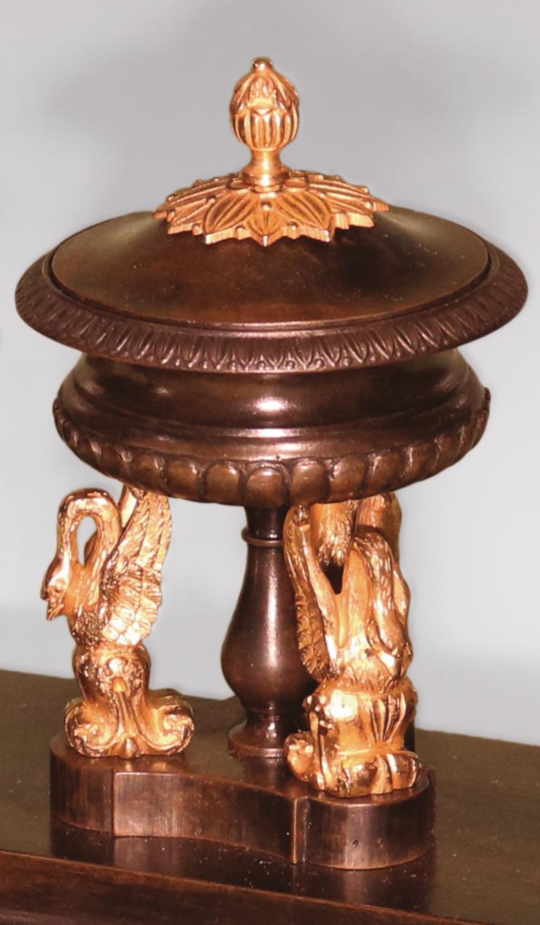 An early 19th century bronze and ormolu pentray, having central covered urn with triple swan mounts, supported on concave triangular plinth base, flanked by leaf and berry decorated campana shaped urns, ending on square block feet.