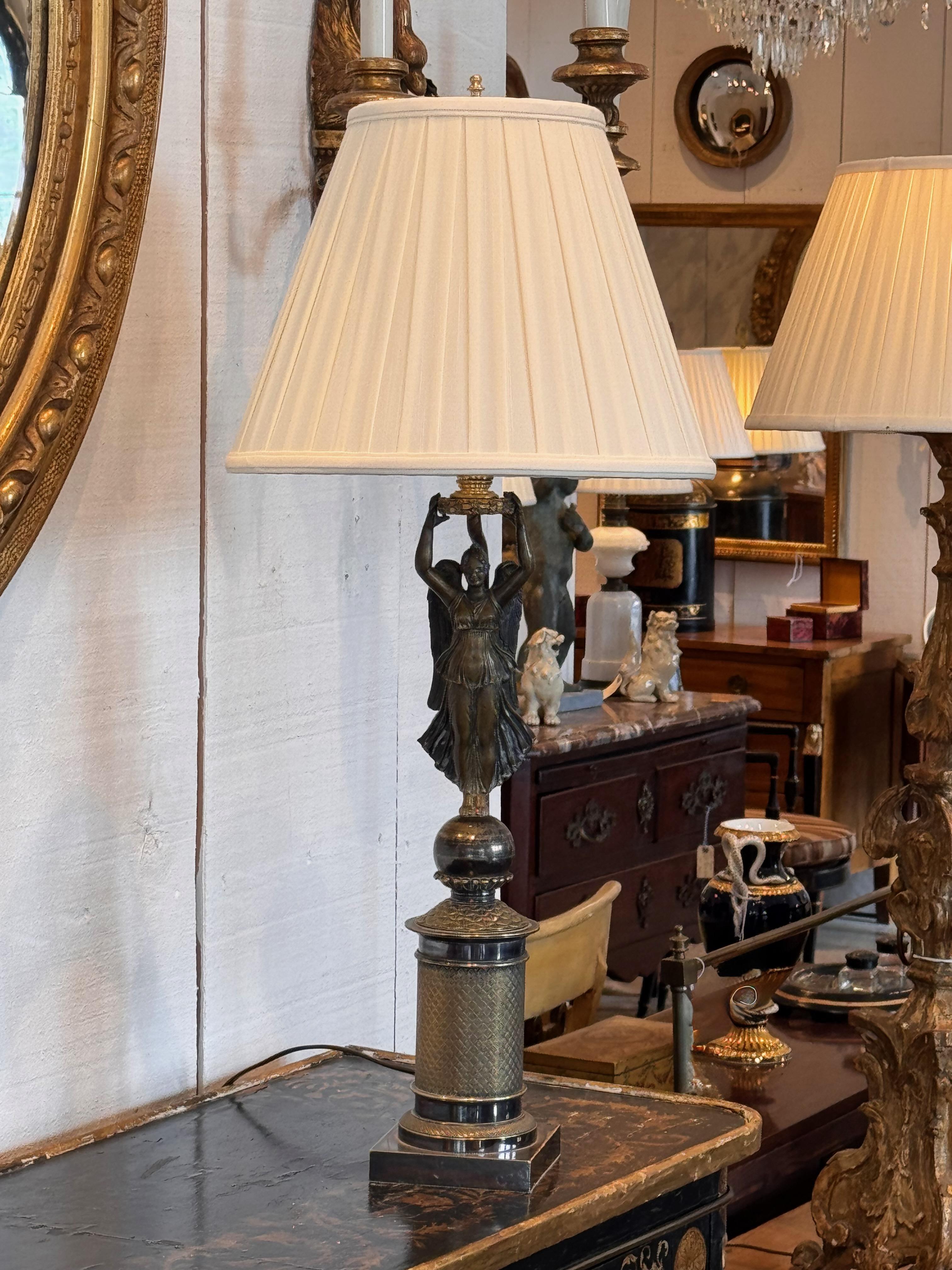 Una lámpara de sobremesa de bronce con un diseño de inspiración neoclásica. La base muestra intrincados detalles con una columna texturizada y acentos decorativos, que le confieren profundidad y carácter. La figura central, que sostiene en alto la