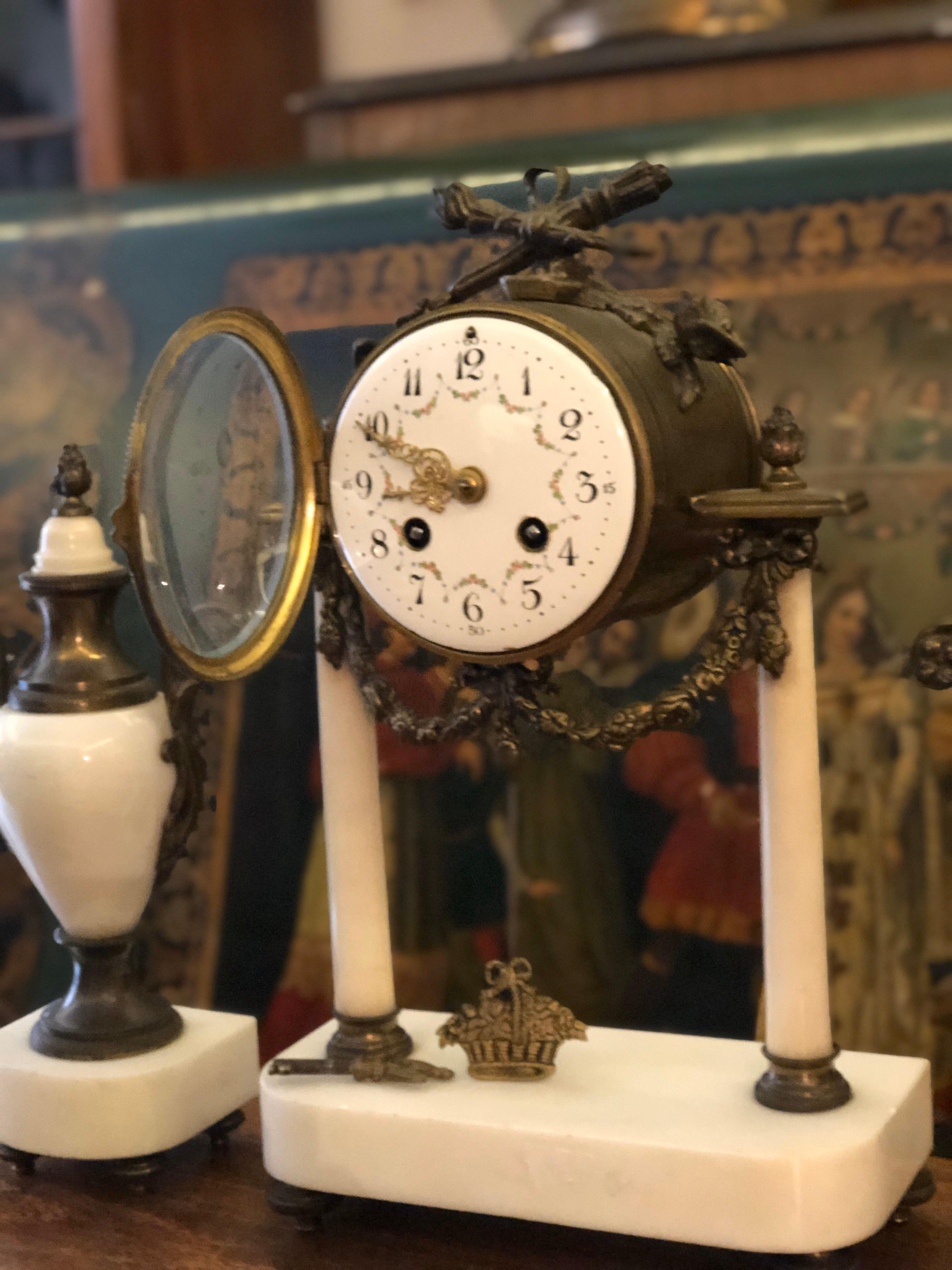 Elegant mantel clock in very good condition with front glass and rich bronze decoration in Louis XVI style, having hand painted porcelain front part and raised on a white marble stand. There are two identical urns surrounding the clock made in the