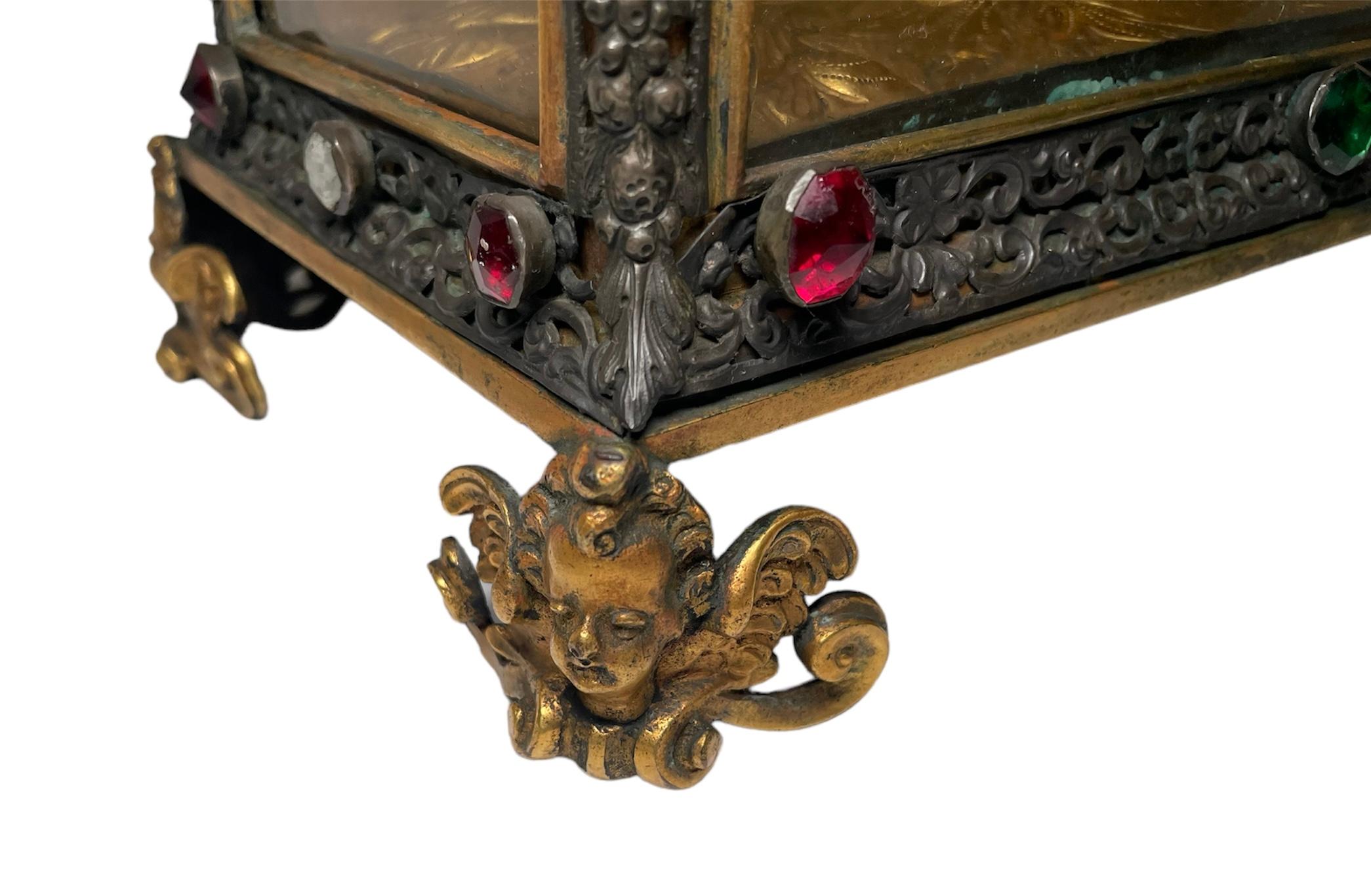 This is a Bronze Reliquary Casket. It depicts a rectangular lidded casket/box   shaped as a house with a hip roof and its walls are glasses framed with patinated bronze. These bronze frames are decorated with a garland of C-scrolls and flowers; and