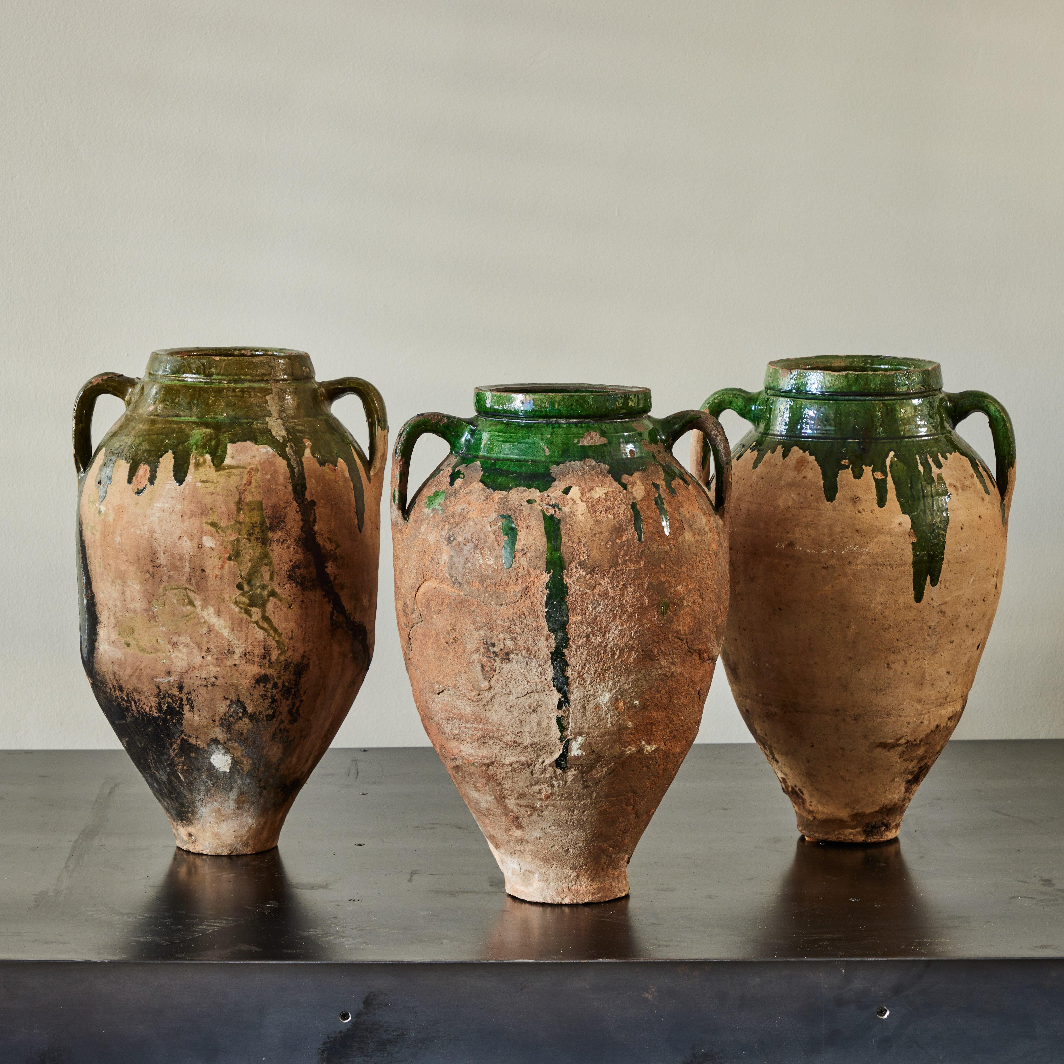 Questi vasi da olio in terracotta bulgara dei primi del Novecento emanano un fascino rustico con le loro forme a mandorla e la loro consistenza invecchiata. Ogni vaso è dotato di due manici sottili ed è impreziosito da uno smalto verde traslucido