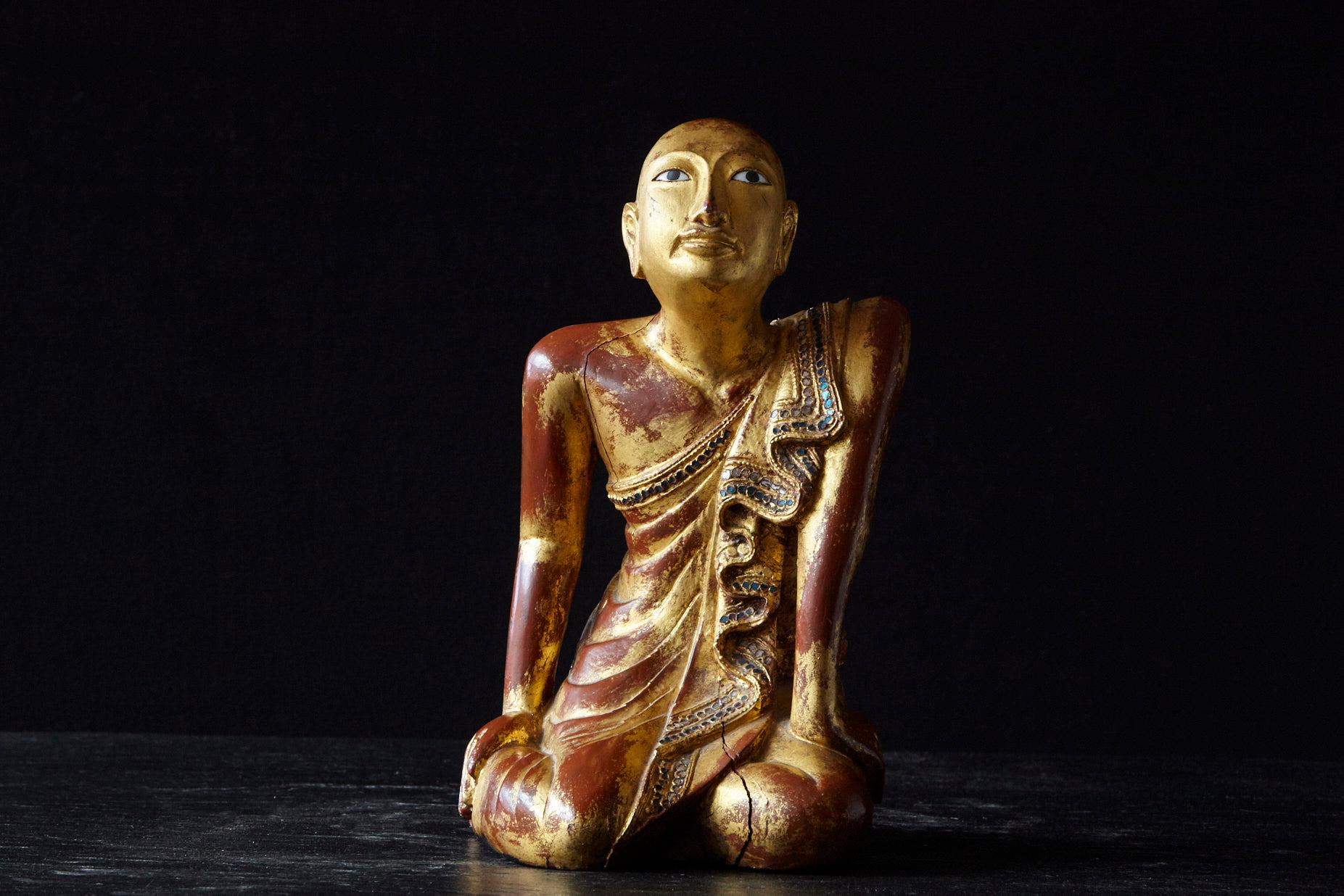 19th century gilded kneeling Burmese Buddhist monk. Carved from one single piece of teak wood, with an underlaying red paint on which the gilt lacquer was applied and with multi colored glass inlays around the edge of the cape of the monk.
The