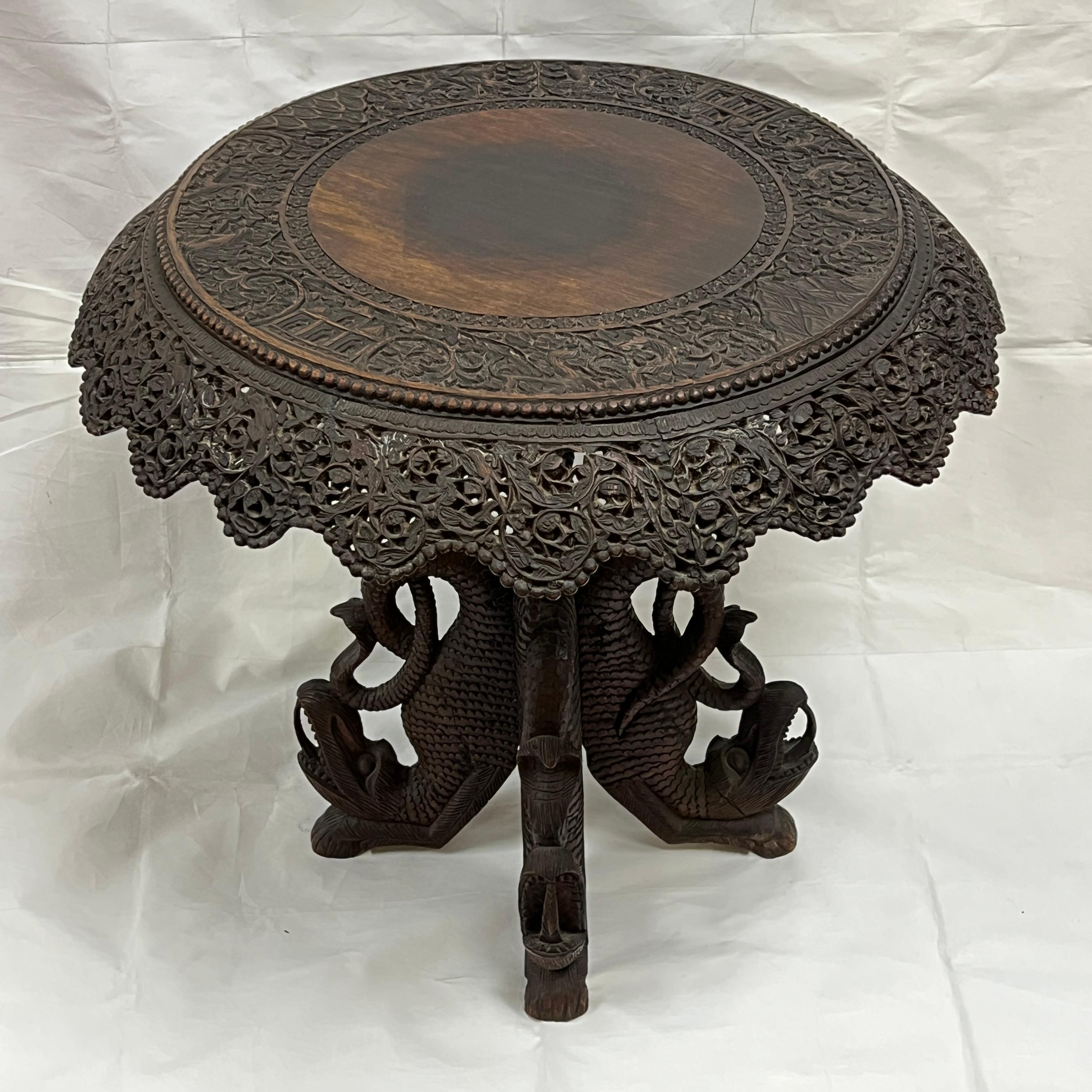 Antique (late 19th century) south asian side table, likely Burmese, 24 inches tall and 26 inches across, featuring finely carved designs of foliage and animals, supported by four legs in the form of dragons and cobra snakes.