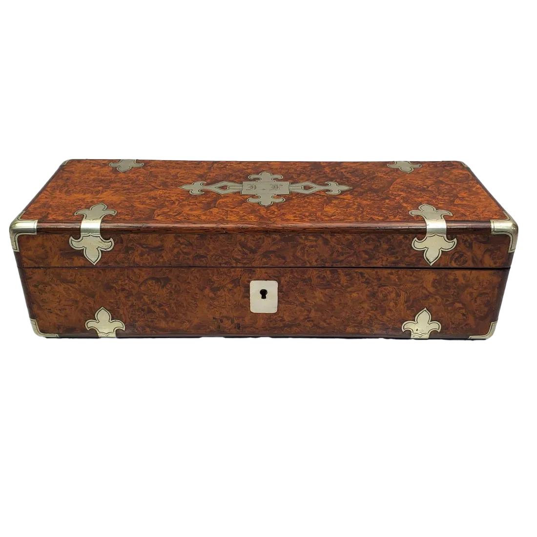 Fina guantera de madera de amboyna burilada Napoleón III con bordes redondeados de nogal y montada con correas de estaño inciso con terminales de flor de lis, las esquinas con monturas conformadas y la tapa centrada con una incrustación metálica que