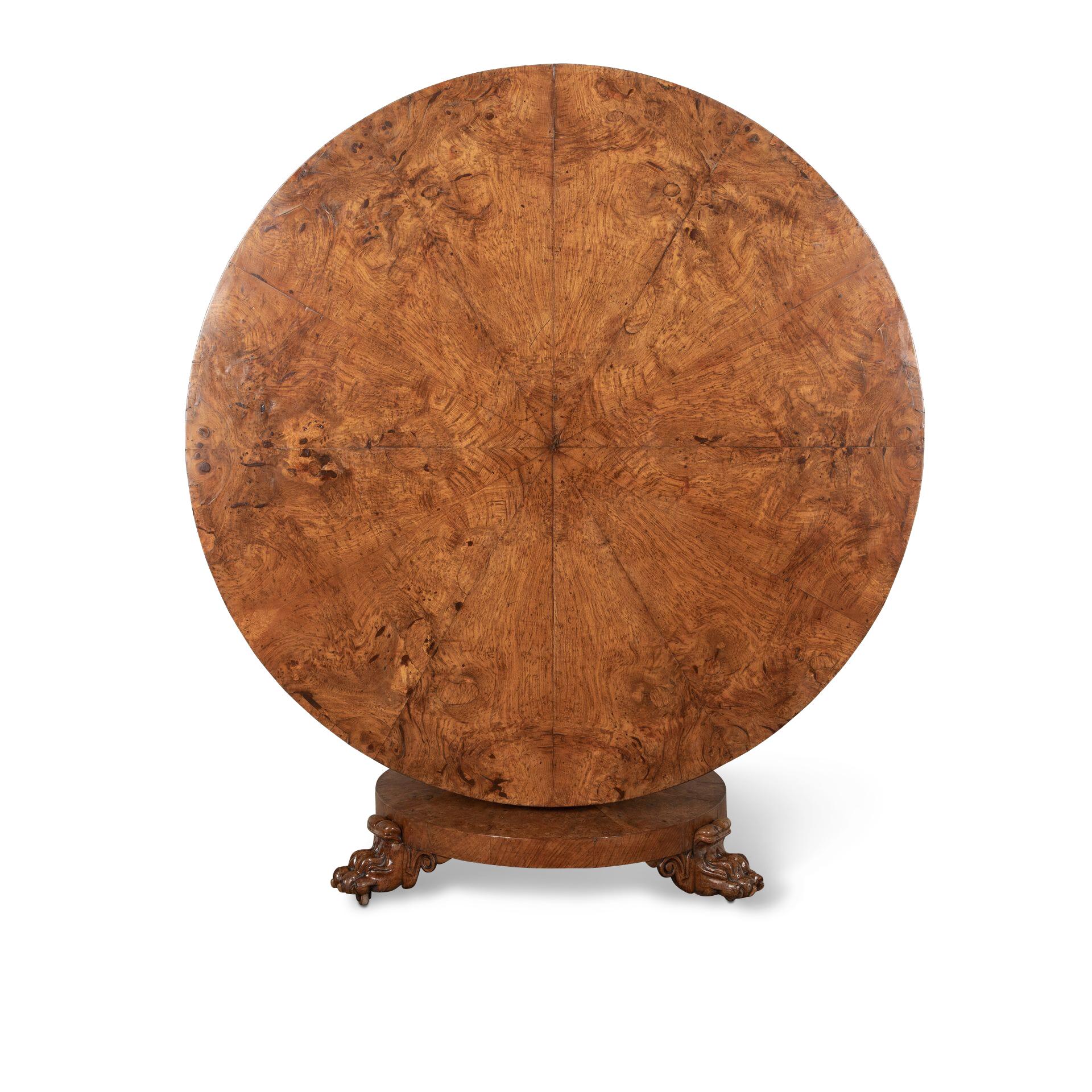 A bold C19th Oak Centre table, the segmented top with well figured burr oak above a shallow frieze, raised on an octagonal column with carved boss and round plinth, raised on carved paw feet flanked with scroll lappets and sunken brass castors.