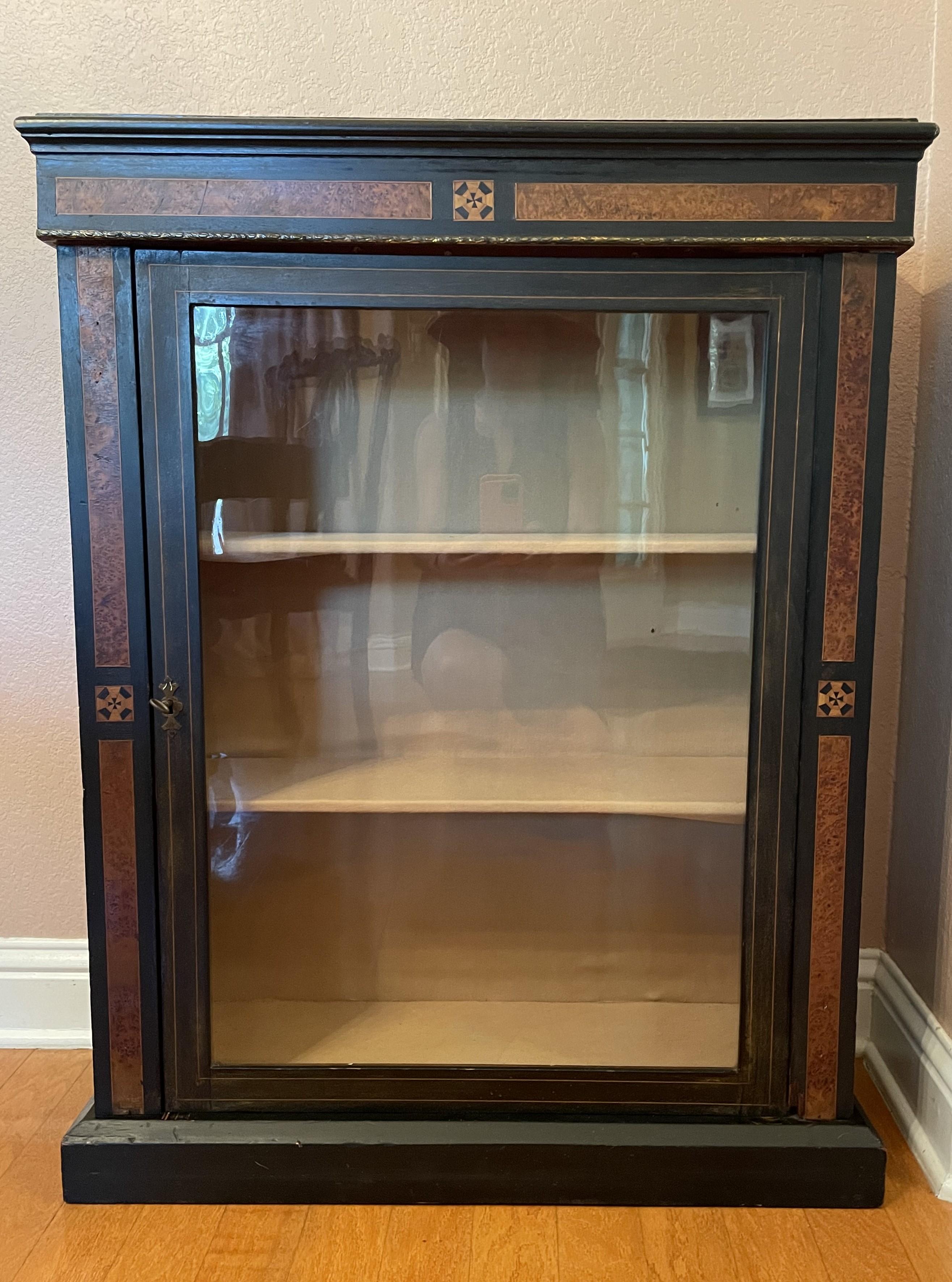 This handsome piece dates to the 19th century. It is most probably a Biedermeier display cabinet, characterized by its structured simple geometry, black wood, burled wood inlays, and antique gold ornamental highlights. The cabinet has three shelves