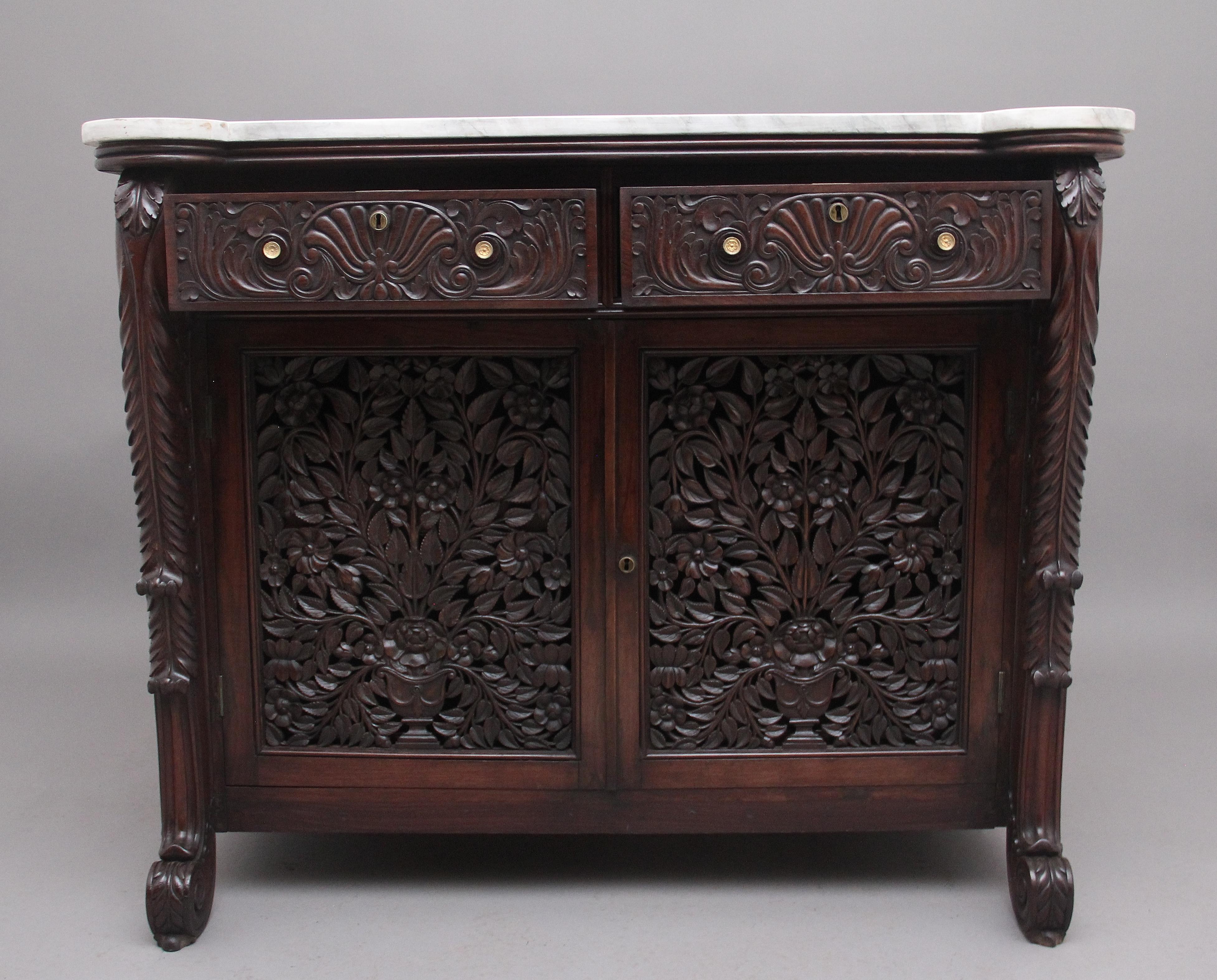 19th century carved Anglo Indian cabinet, having the original white veined shaped marble top above two frieze drawers with the original turned brass engraved handles, the drawer fronts having carved floral and shell decoration, two hinged cupboard
