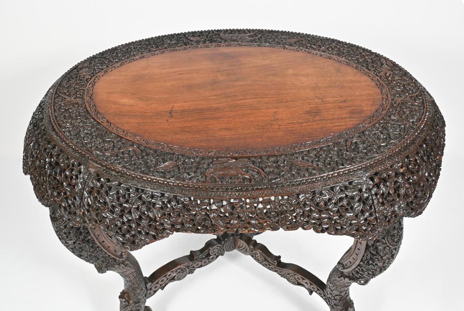 An elaborately carved center table, oval shaped with a carved surface and beaded trim. Having a profusely carved and pierced foliate vine motif apron. The carvings extend down the four scroll legs, all joined by an x-form stretcher with bird form