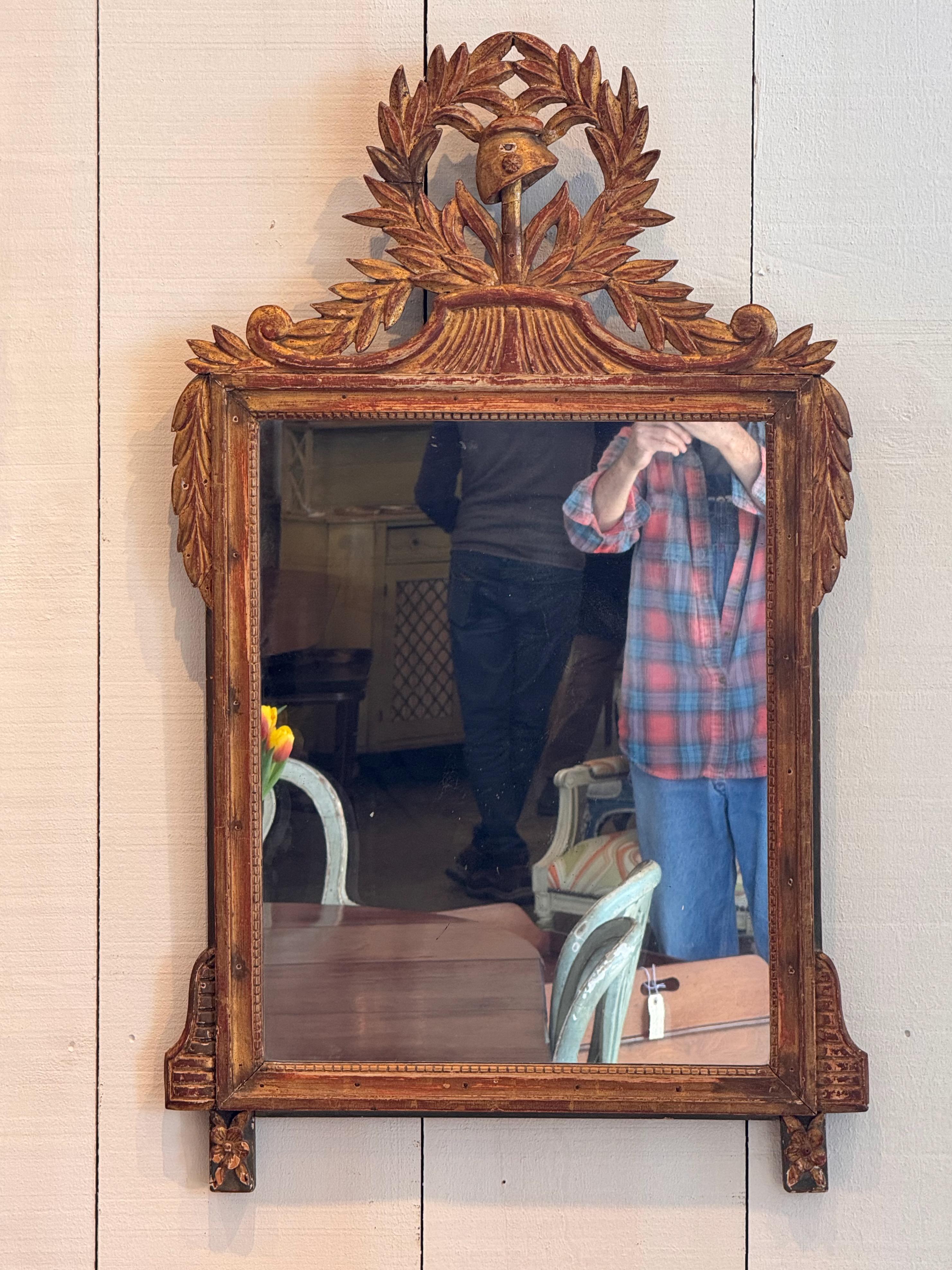 Elegant miroir en bois doré du 19ème siècle avec des détails complexes sculptés à la main. Le cimier représente une couronne de laurier entourant un casque, symbole de la victoire et de la force. Le cadre est orné de motifs classiques, notamment de