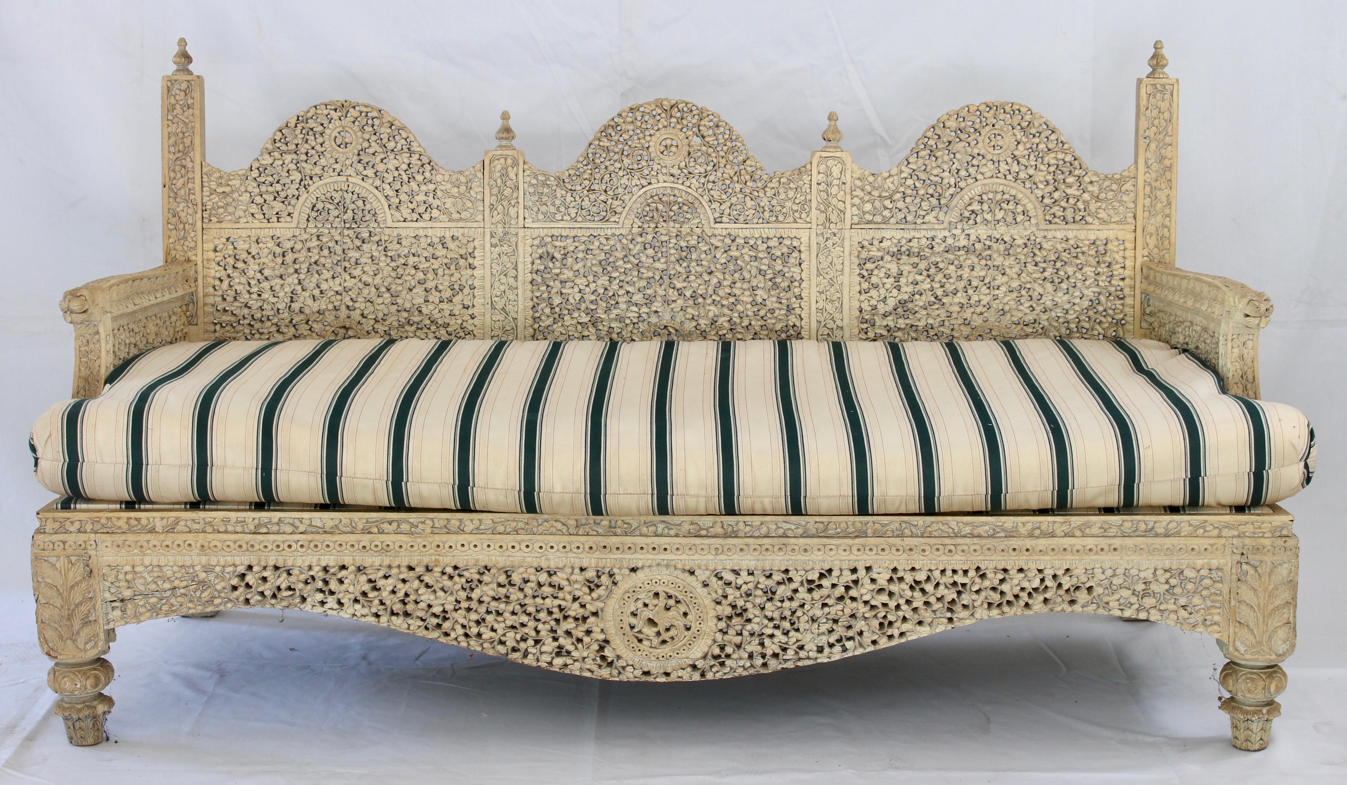 An exceptional heavily carved Indian sofa or settee with original cream colored paint finish and likely later upholstered cushion in green and ivory striped cotton fabric.