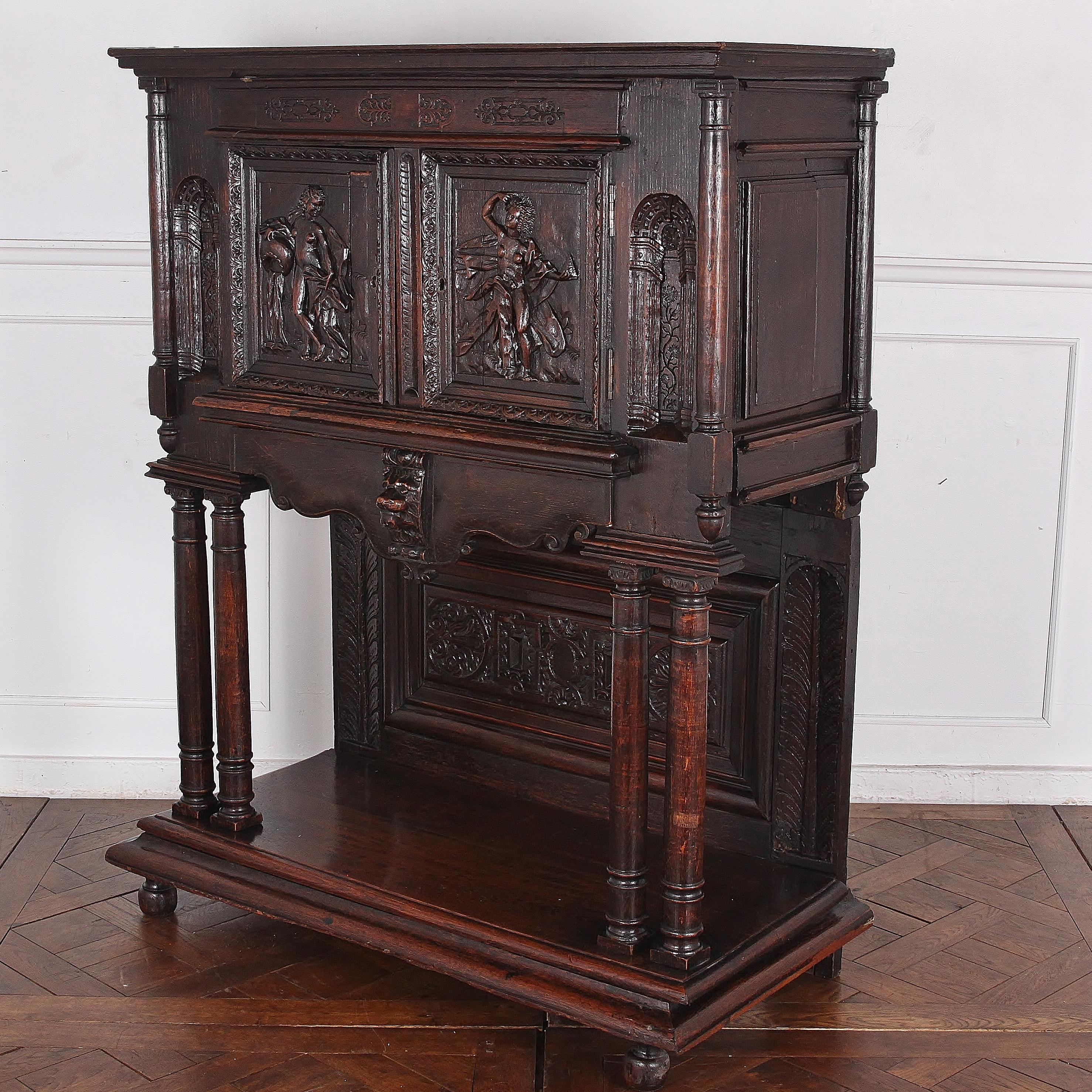 A highly-carved oak Renaissance-revival cabinet on stand, the top with figural carving to the doors, the doors flanked with trompe l’oiel carved arcades, the whole raised on double-column supports over a boldly-molded lower shelf with turned bun