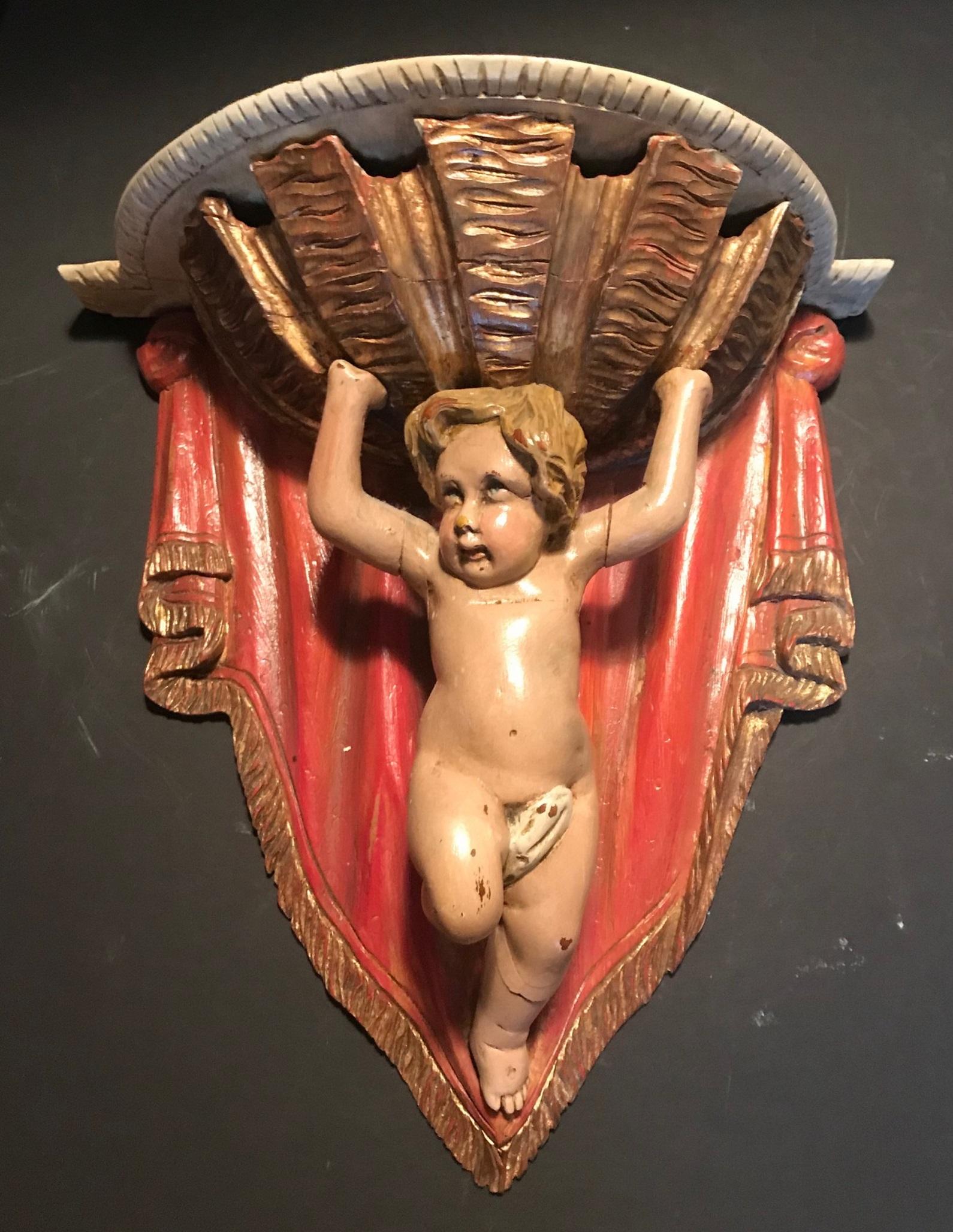 The little putto is supporting a big gilded basket against a red drape. This 19th century French carving is done in great detail. The polychrome decoration is painted in vibrant colors. It is a charming and decorative wall hanging shelf with