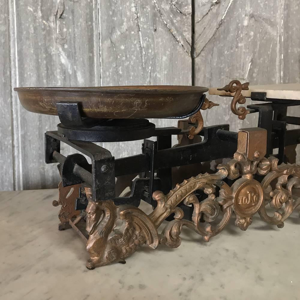 19th century cast iron balance scale features an elaborate scrolled design with a marble platform on one side of the scale, and a shallow brass bowl on the other. Gold painted highlights indicate this example was placed in a position of prominence