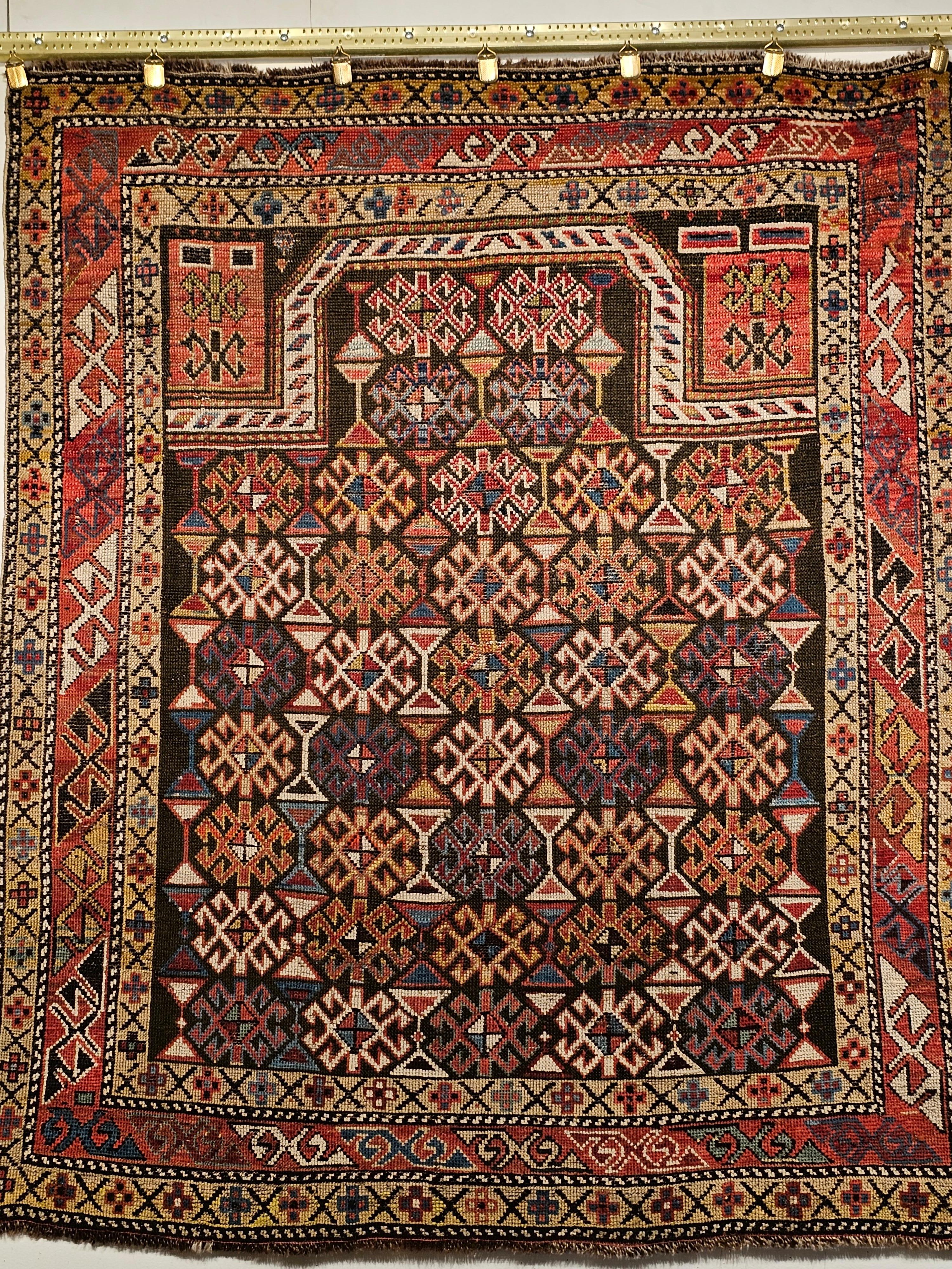 A beautiful Caucasian Shirvan area rug from the southern Azerbaijan region of the Caucasus is in a prayer rug design with an abrash dark brown field.  The field design elements include the “hour glass” and the star patterns repeated in each row. 