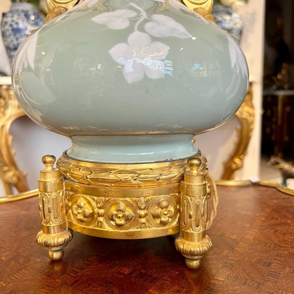 This large celadon porcelain lamp, measuring 50 cm in height and 22 cm in diameter, showcases intricate floral and vegetal motifs, a creation from the esteemed French workshop Gagueneau. It is elegantly adorned with a chiselled and gilded bronze