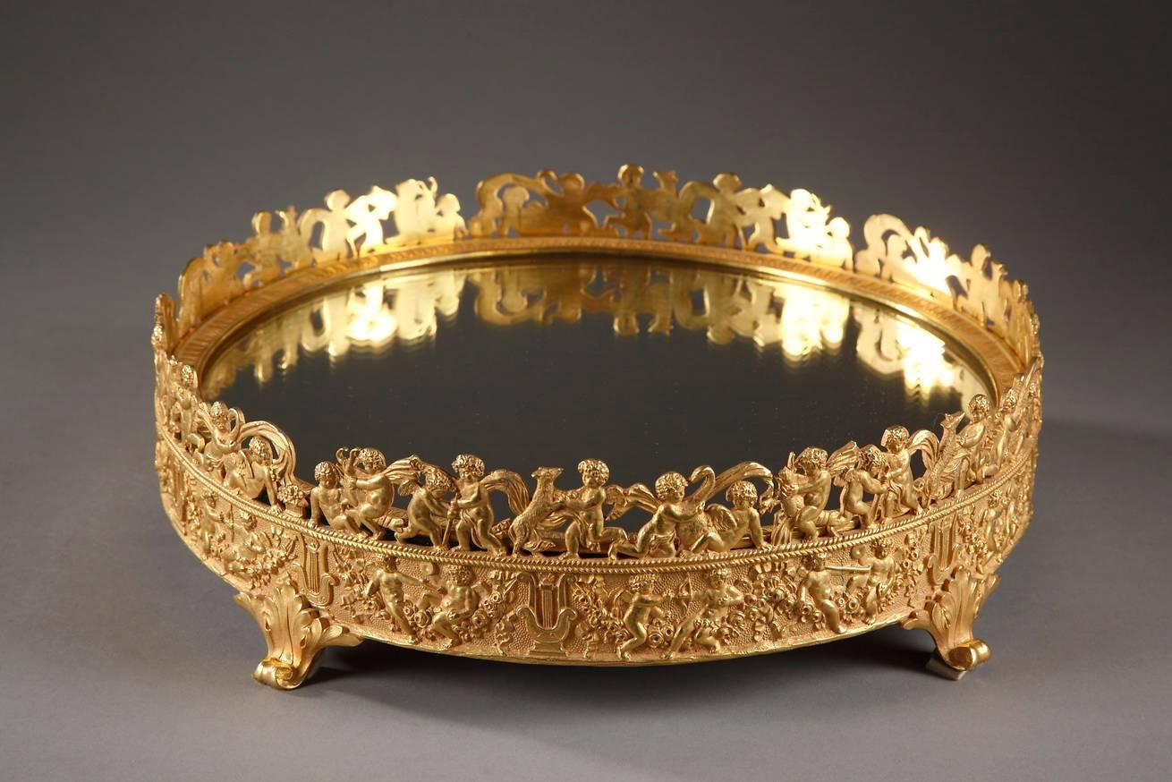 Circular gilt and chiseled bronze centerpiece with four foliated feet. The circular base is covered with a mirror and is surrounded by two friezes of cupids wearing sashes. On the lower frieze, the cupids are playing music and drawing their bows