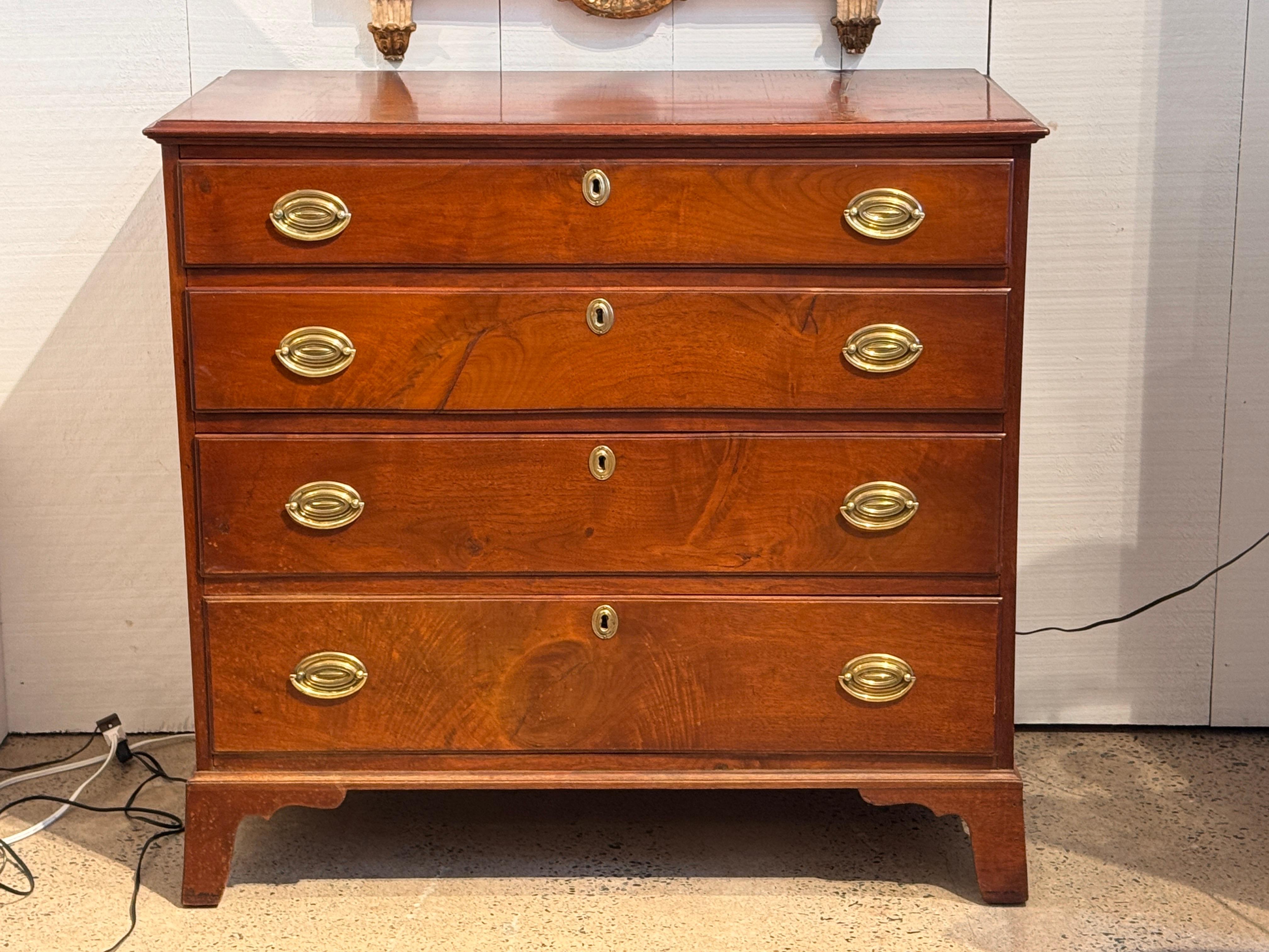 Cómoda del siglo XIX con cuatro cajones graduados, cada uno de ellos provisto de tiradores y escudos ovalados de latón. La parte superior tiene un borde moldeado, y la caja se apoya en pies de ménsula perfilados. La construcción de los cajones