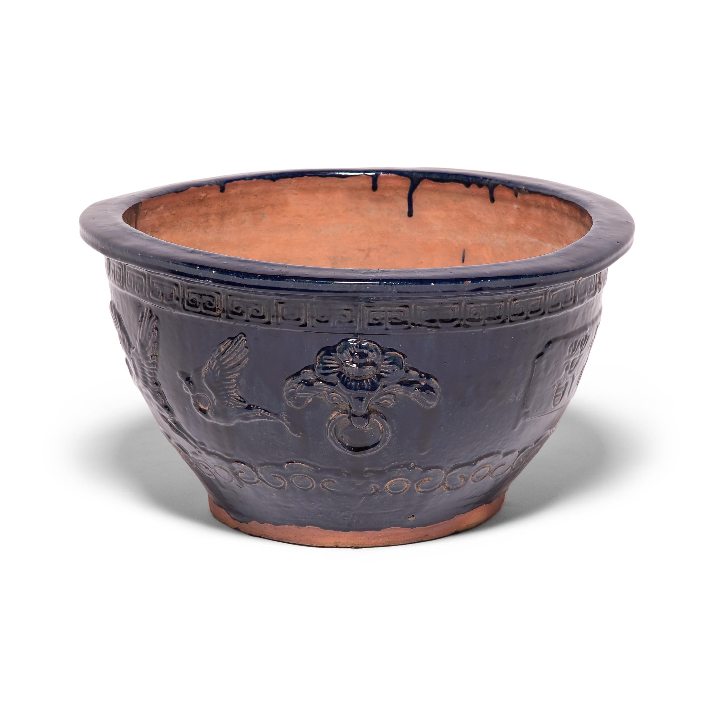 This shallow terracotta bowl is delicately decorated in low relief and cloaked in a monochrome layer of dark blue glaze. Framed by a Meander trim and billowing cloud motifs below, two cranes fly towards a pine tree with wings outspread. In