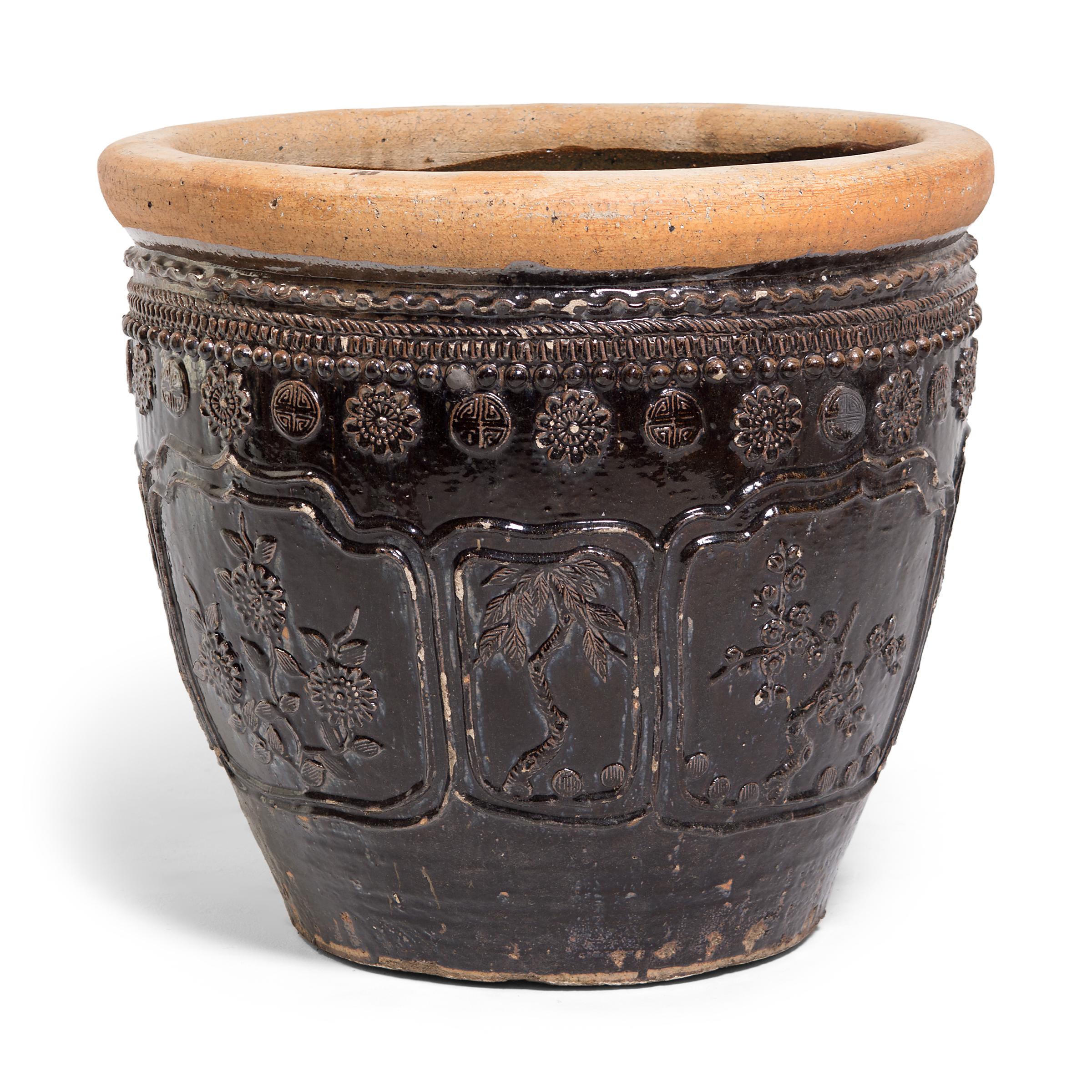 This large glazed jar was handcrafted in China's Shanxi province over a century ago. Masterfully crafted, the jar is patterned in high relief, featuring medallion portraits of chrysanthemums, plum blossoms, bamboo, and pine branches. A textured trim