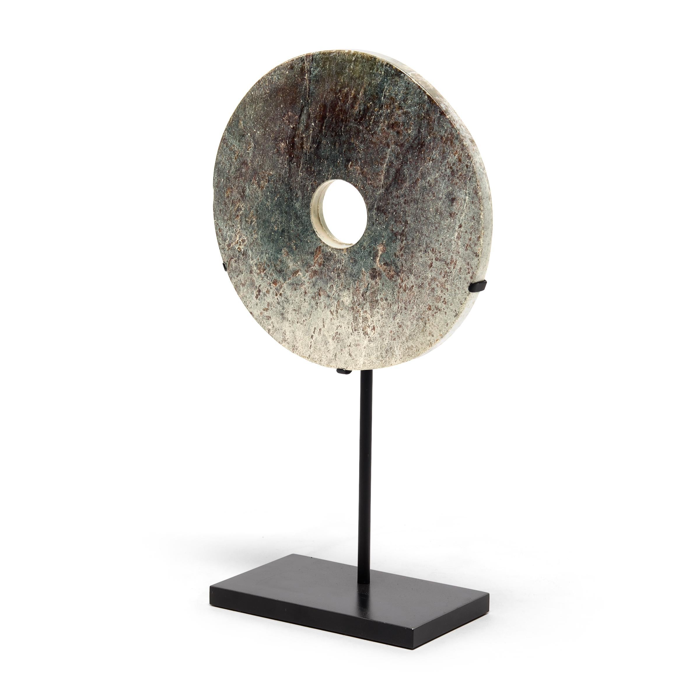 This petite bi disc is softened by occlusions of grey and purple that contrast the dark stone like an encroaching winter's Frost. Found in the tombs of ancient Chinese emperors and aristocrats, bi discs such as this have a mysterious and spiritual