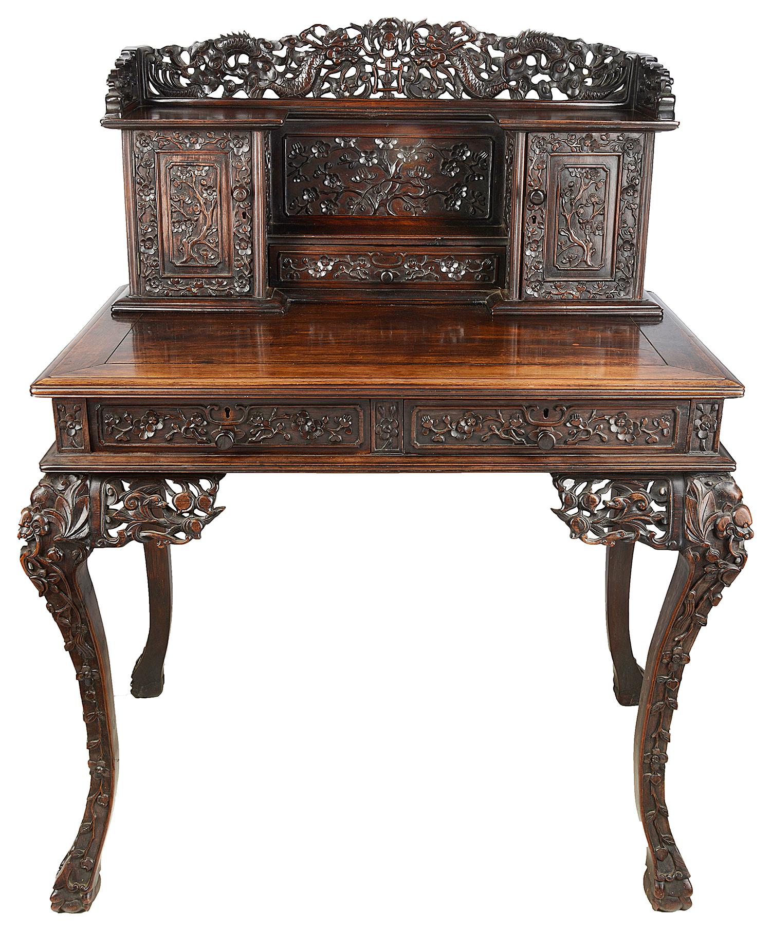 A good quality 19th century Chinese hardwood writing desk, having wonderful hand carved mythical dragons to the gallery, two cupboard doors and drawer all with carved blossom tree decoration, a polished writing surface, two frieze drawers, raised