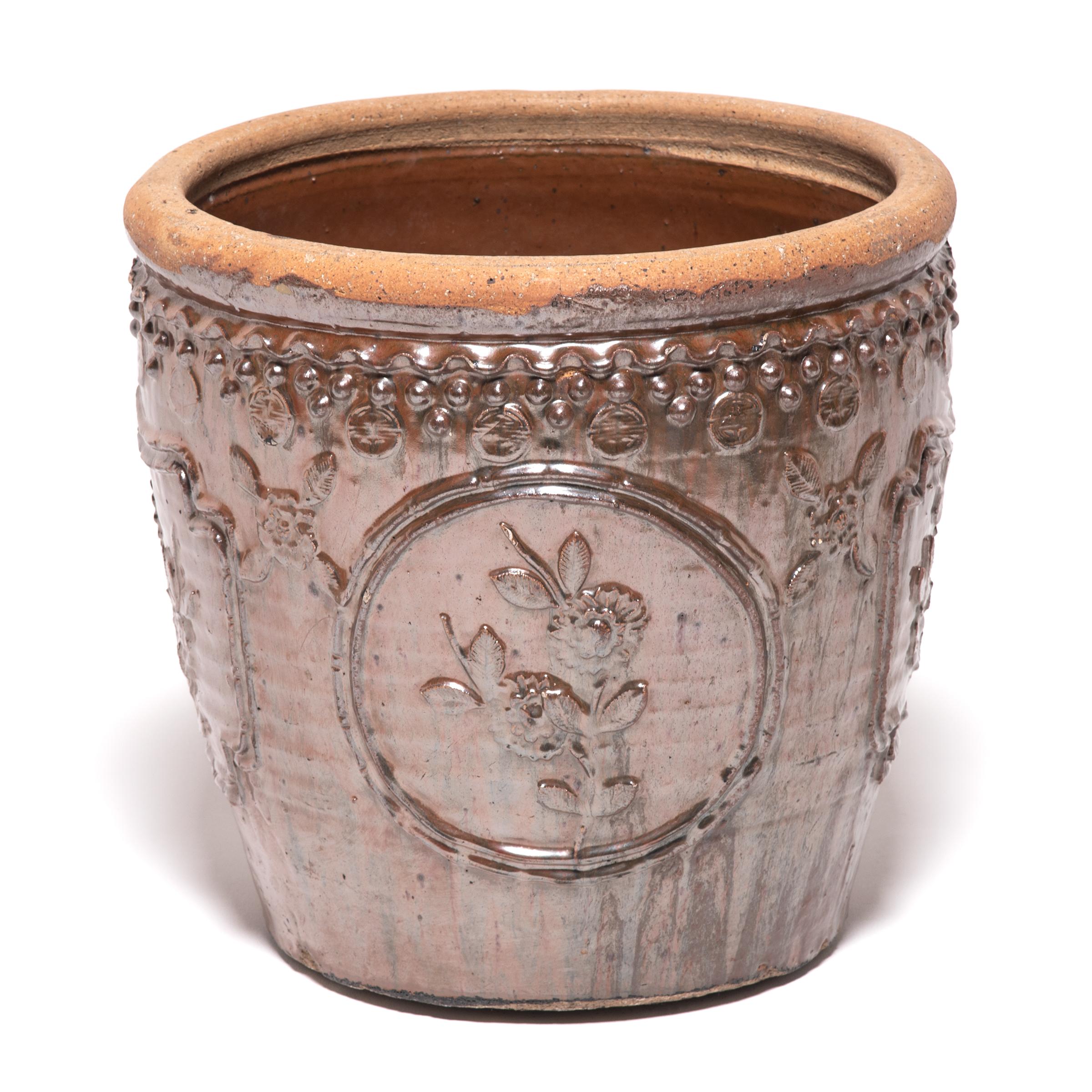 This large glazed jar was handcrafted in Northern China over a century ago. Masterfully crafted, the jar is patterned in high relief featuring medallion portraits of blooming chrysanthemums and trimmed around the rim with bead-like motifs.