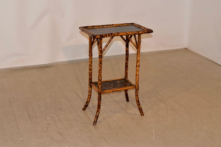 19th century tortoise bamboo side table from France with a chinoiserie decorated top, supported on legs, which are connected by a lower shelf. The shelf is covered with a painted wallpaper. The legs end in splayed feet.