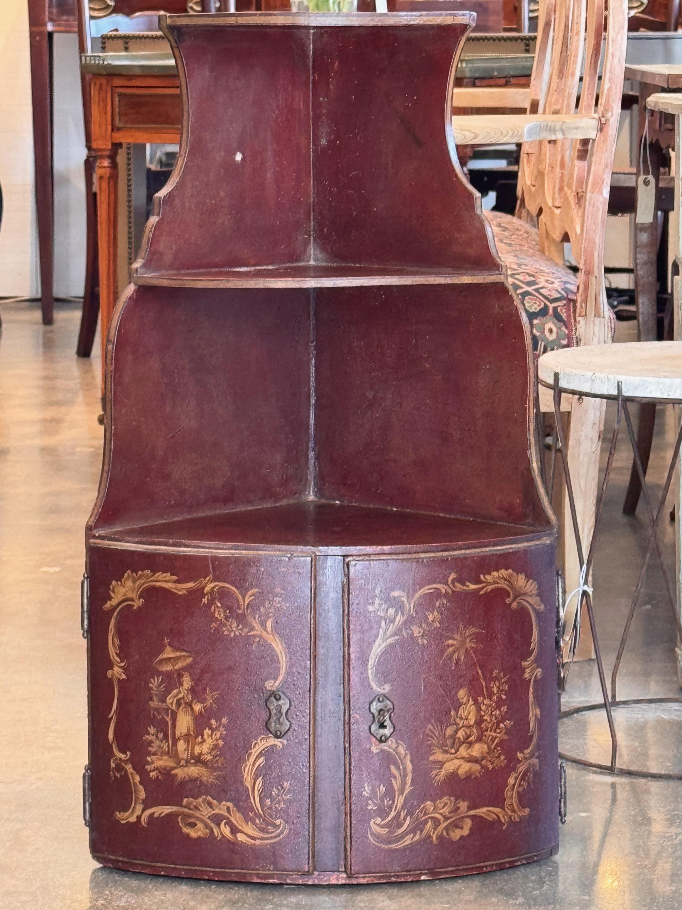Questo mobile angolare Chinoiserie del XIX secolo presenta una ricca finitura in lacca rossa con delicate scene dipinte in oro. La parte superiore è dotata di scaffali curvi, mentre lo scomparto inferiore offre un vano portaoggetti dietro due ante