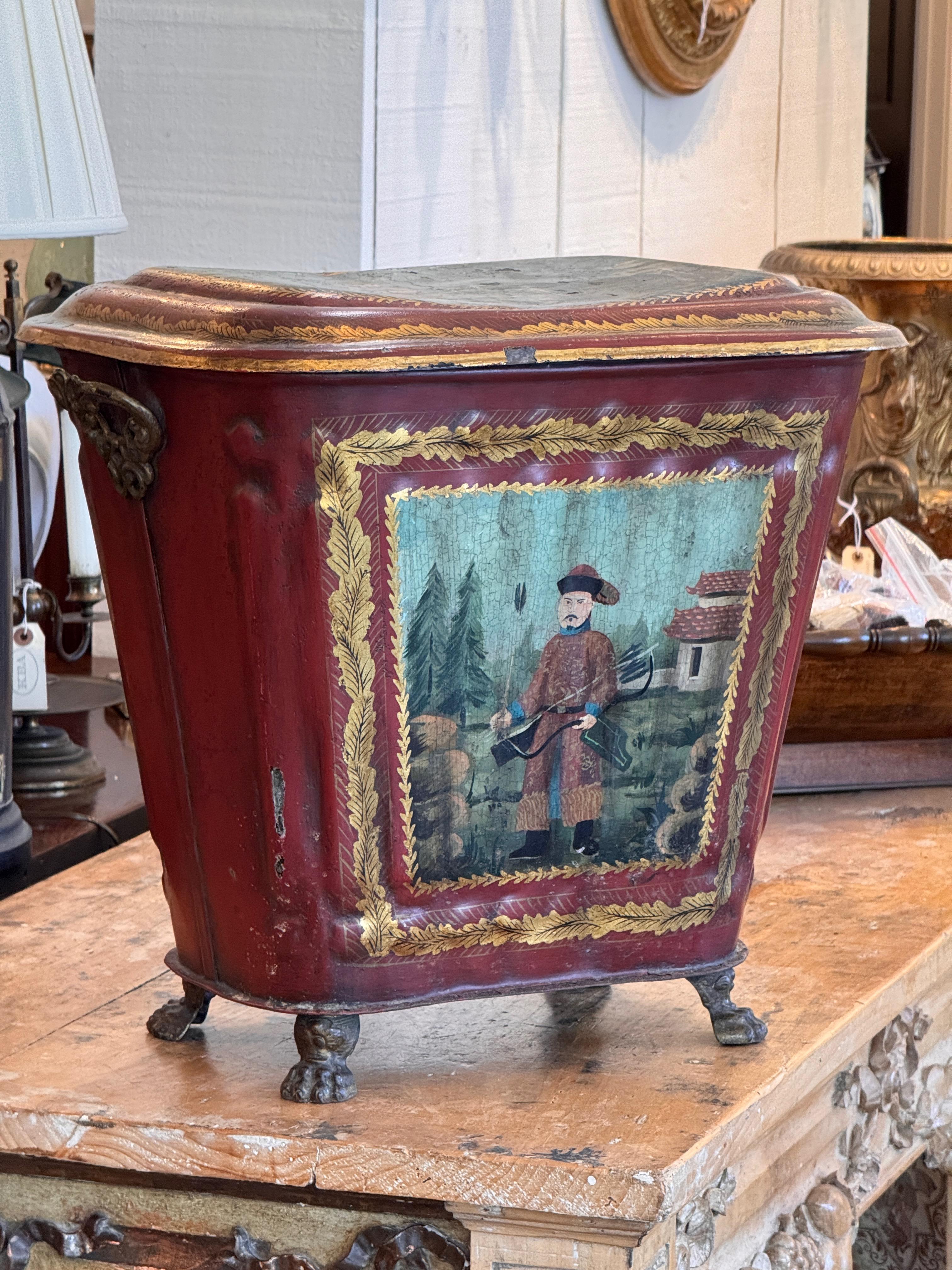 Corbeille à charbon décorative en tole présentant des scènes de Chinoiserie peintes à la main et encadrées de bordures feuillagées dorées. Le corps peint en rouge repose sur des pieds en forme de pattes et comprend des supports latéraux en laiton.