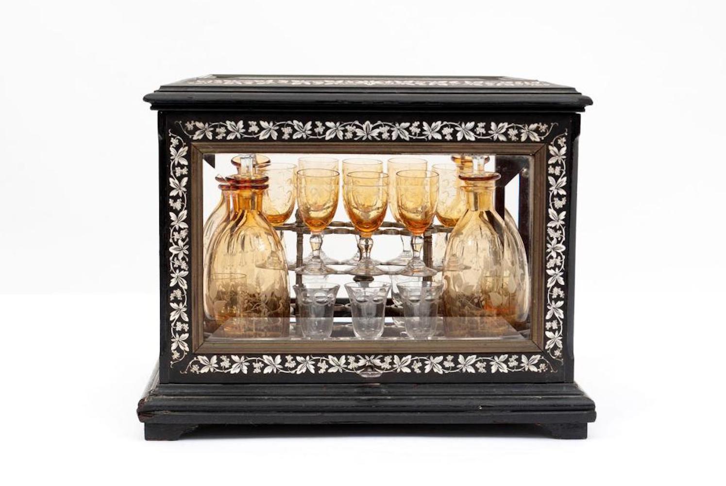 Cave a liqueur.
Continental, 19th century. Ebonized wooden case with all around beveled glass panels with hinged front to top panels, fitted with a full Tantalus rack of etched amber crystal decanters and diminutive liqueur glasses. Measures: 11