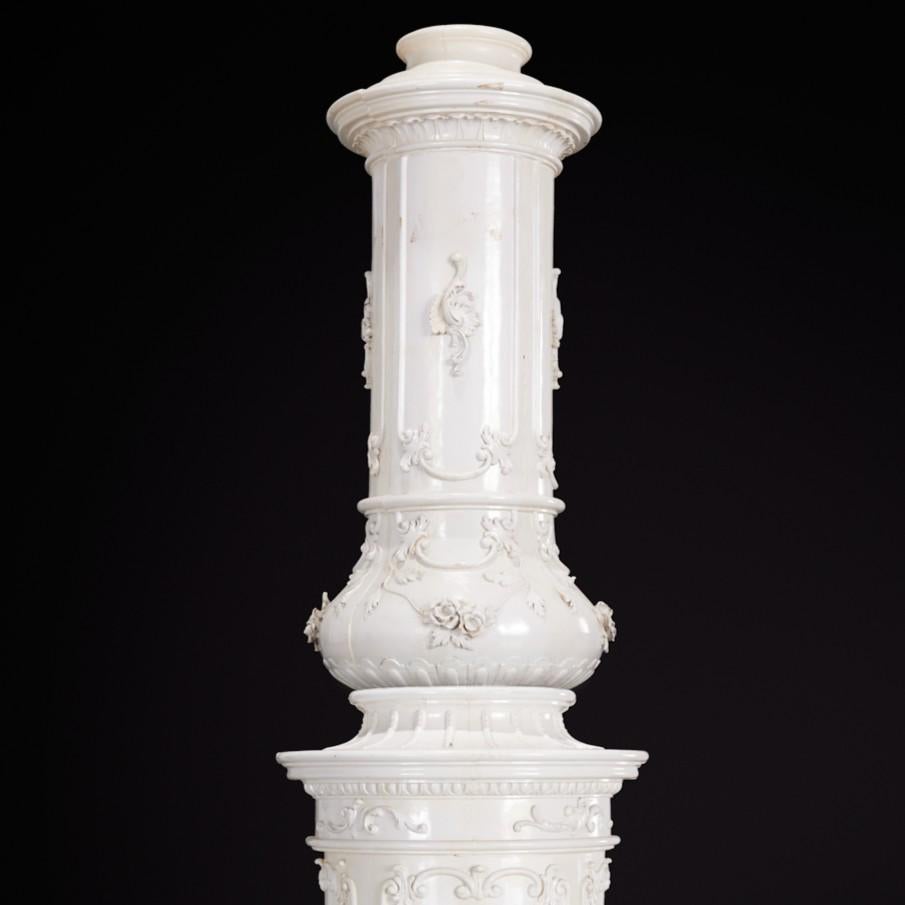 France or Italy, 19th Century, tin-glazed terracotta, in three stacked sections with domed wooden cap, featuring raised foliate scrollwork, floral garlands, and Rococo-inspired cartouches, all in a cream-white glaze. Brass fire and ash doors with