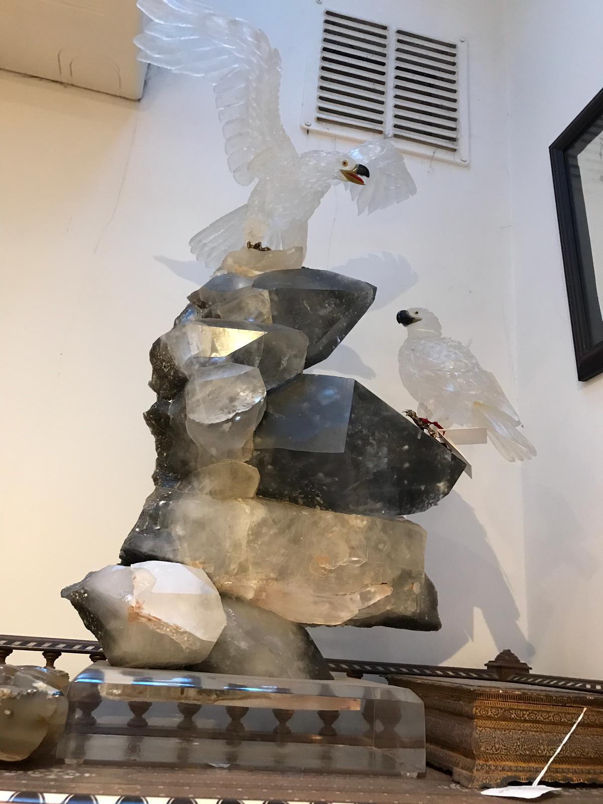 It is a rock crystal with gold and precious stone featuring both its natural geometric formation on a square cut base, along with a pair of skilfully cut eagles sitting upon it. One is resting peacefully to the right, whilst the other is in movement
