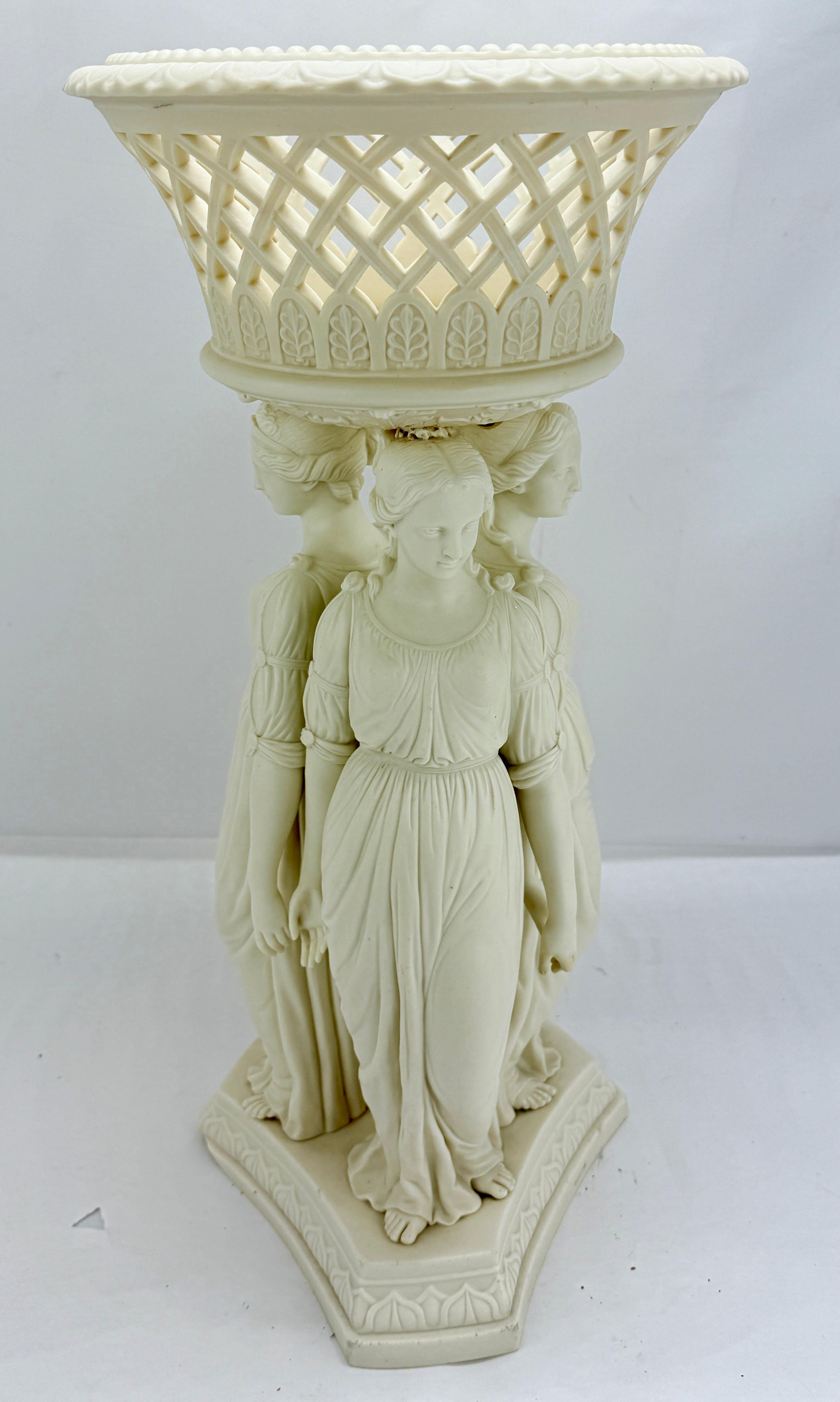 Il centrotavola è modellato interamente in Parian color crema con una lucentezza morbida e satinata che ricorda il marmo finemente cerato. Tre fanciulle a figura intera stanno spalla a spalla su un basamento a trifoglio, con il peso leggermente