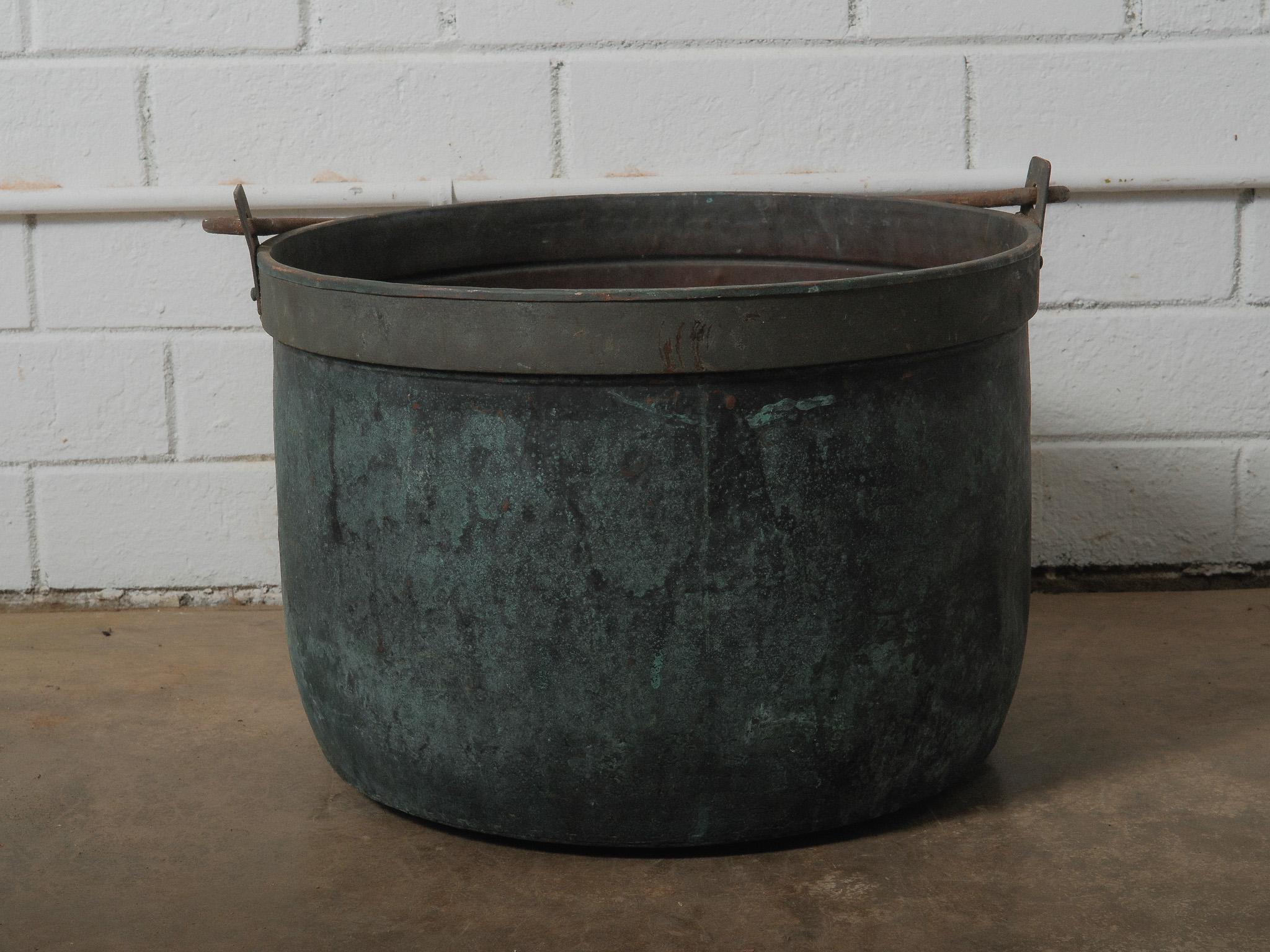 A substantial hand-formed copper vessel with dramatically aged green patina, presenting a deep cylindrical body and fitted with iron swing handles set into riveted brackets. The silhouette is defined by its weighted proportions, upright stance, and