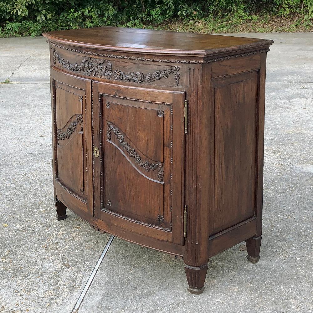 19th century Country French fruitwood buffet features an unusual subtle bowfront facade, festooned with classical bas relief that would please King Louis XVI and his queen, Marie Antoinette! Ribbon, wildflowers and foliates arranged in swags and